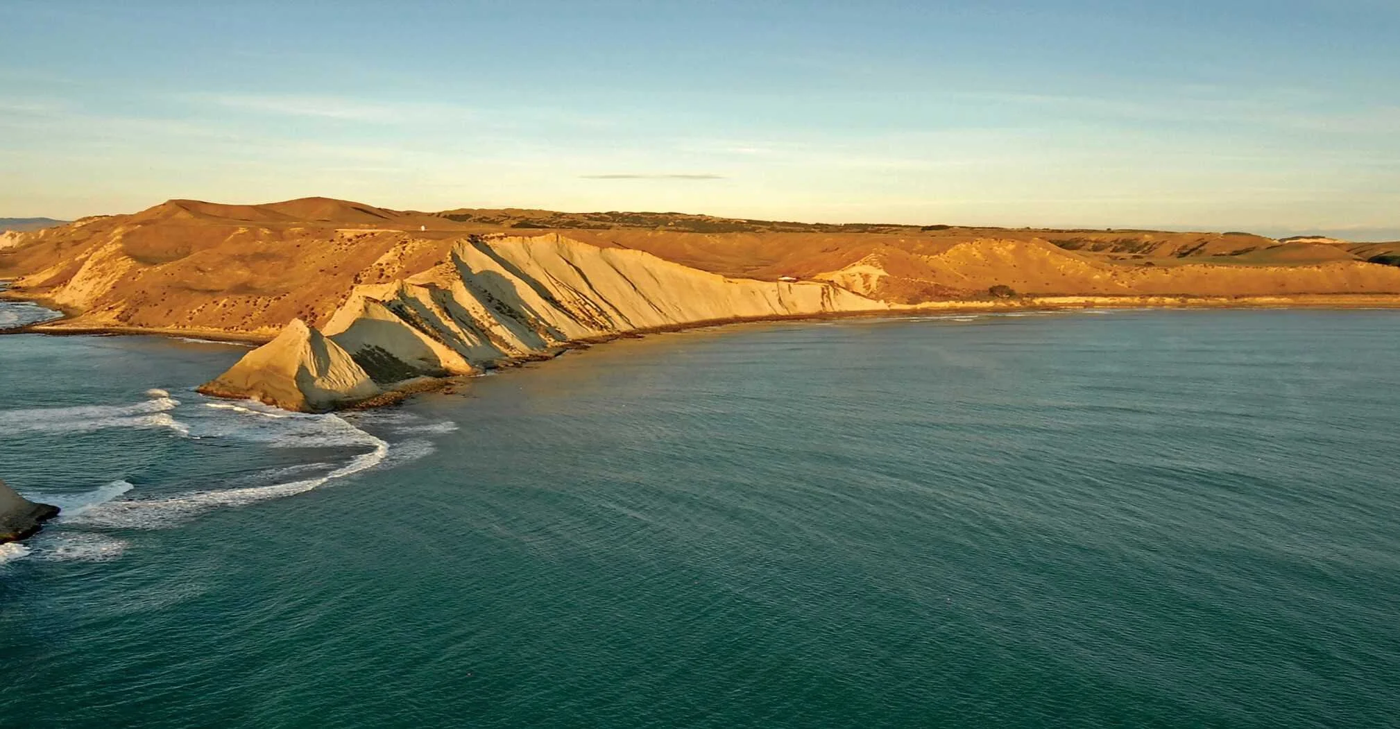 Aerial view of white cliffs along a coastline with the ocean in the foreground and rolling hills in the background during sunset.
