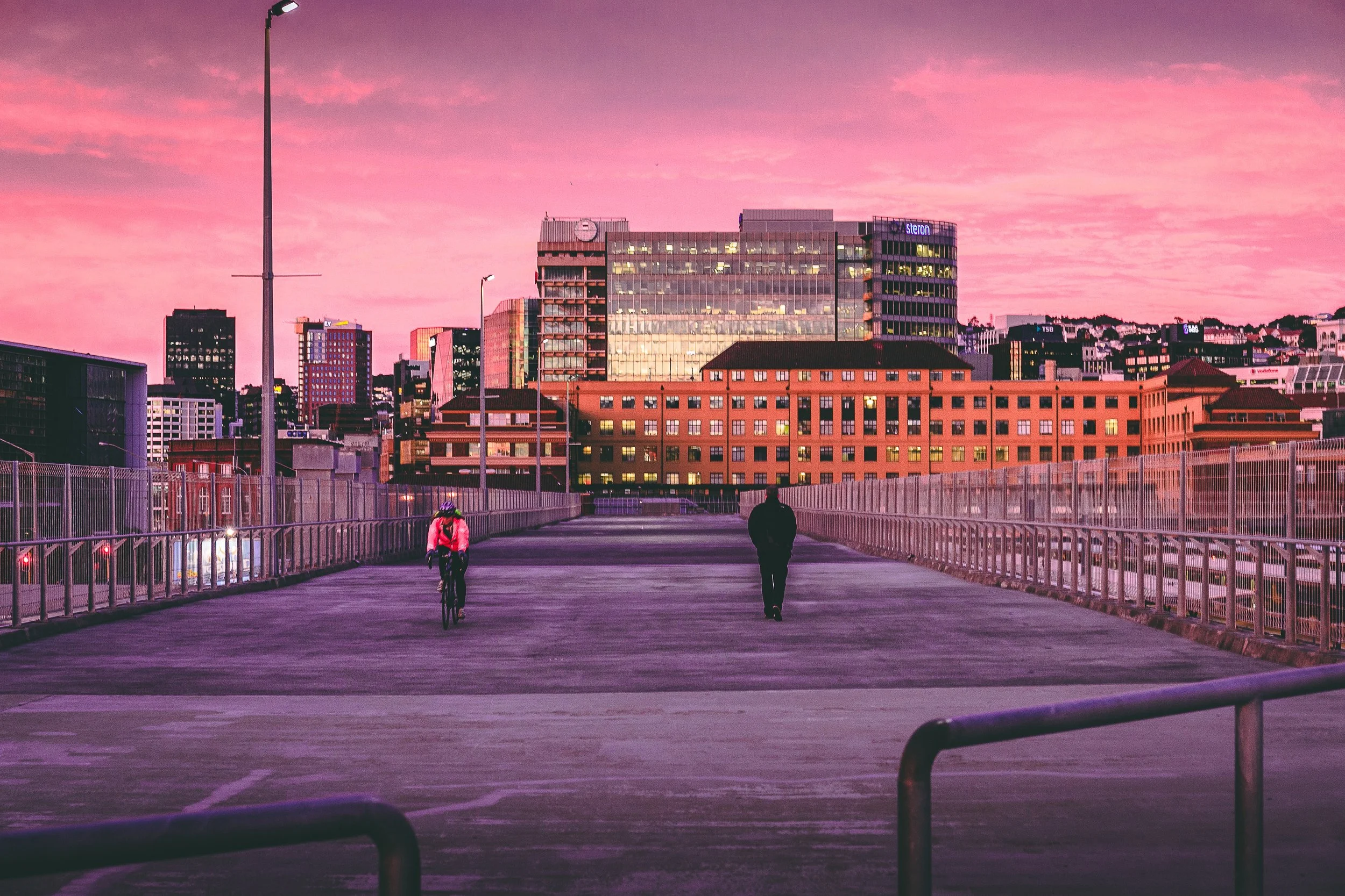 Cityscape at sunset with pink and purple sky, tall buildings with illuminated windows, and a bridge or walkway in the foreground where a cyclist and a pedestrian are walking.