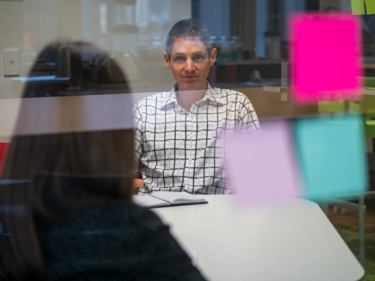 Two people having a discussion in an office, seen through a glass wall with colorful sticky notes on it. One person is sitting at a desk, the other is sitting opposite, with an open notebook in front of him.