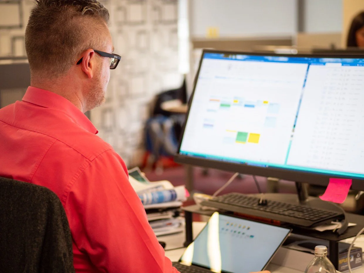Man wearing glasses in a red shirt working at a desk with a large computer monitor displaying a calendar or scheduling app and a tablet.