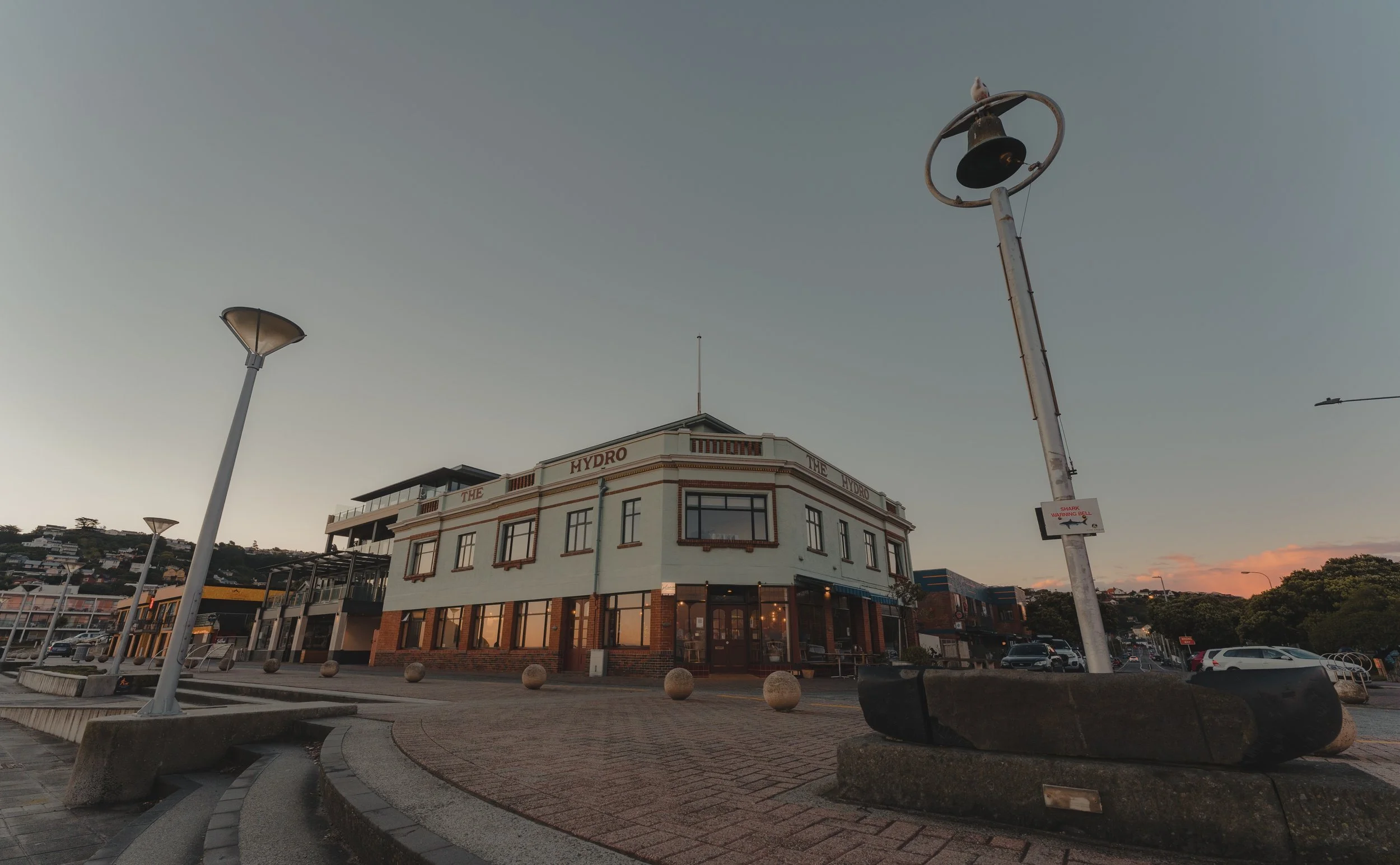 A corner of a city street at dusk with a large white building labeled 'THE HYDRO'. There are multiple street lamps, one with a bird perched on top, and a sign warning about a shark warning bell. Cars are parked along the street, and there are houses 