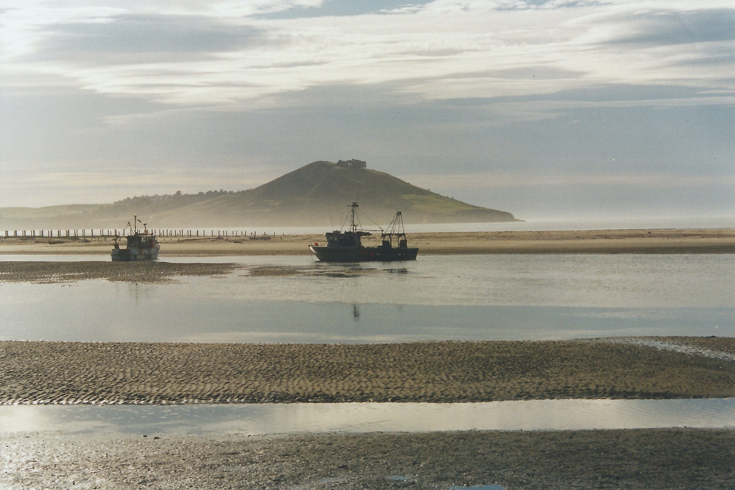 Two boats floating in calm water near a sandy shoreline, with a hill and cloudy sky in the background.