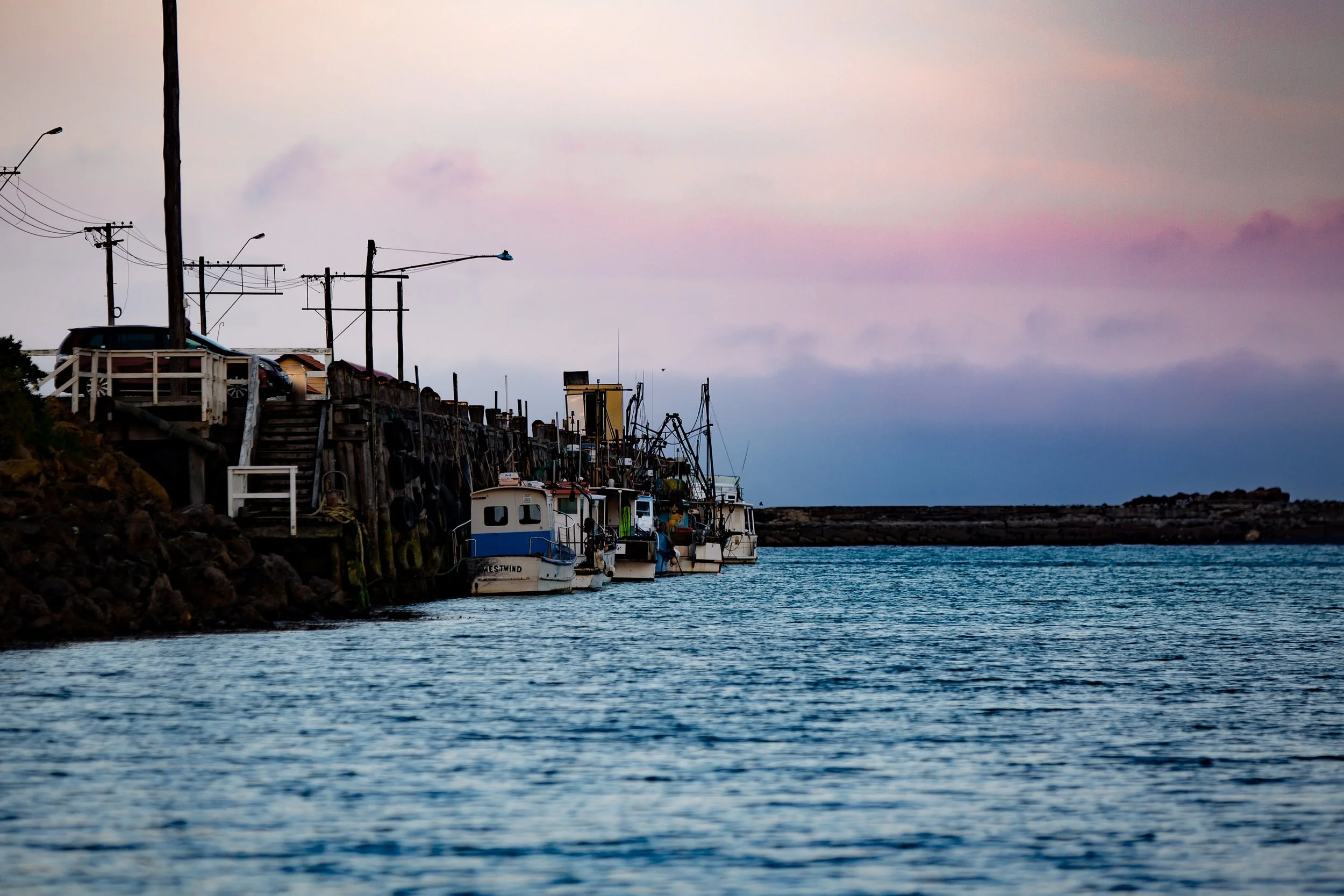 A harbor scene during dusk with boats docked along a wooden pier and power lines against a pastel-colored sky.