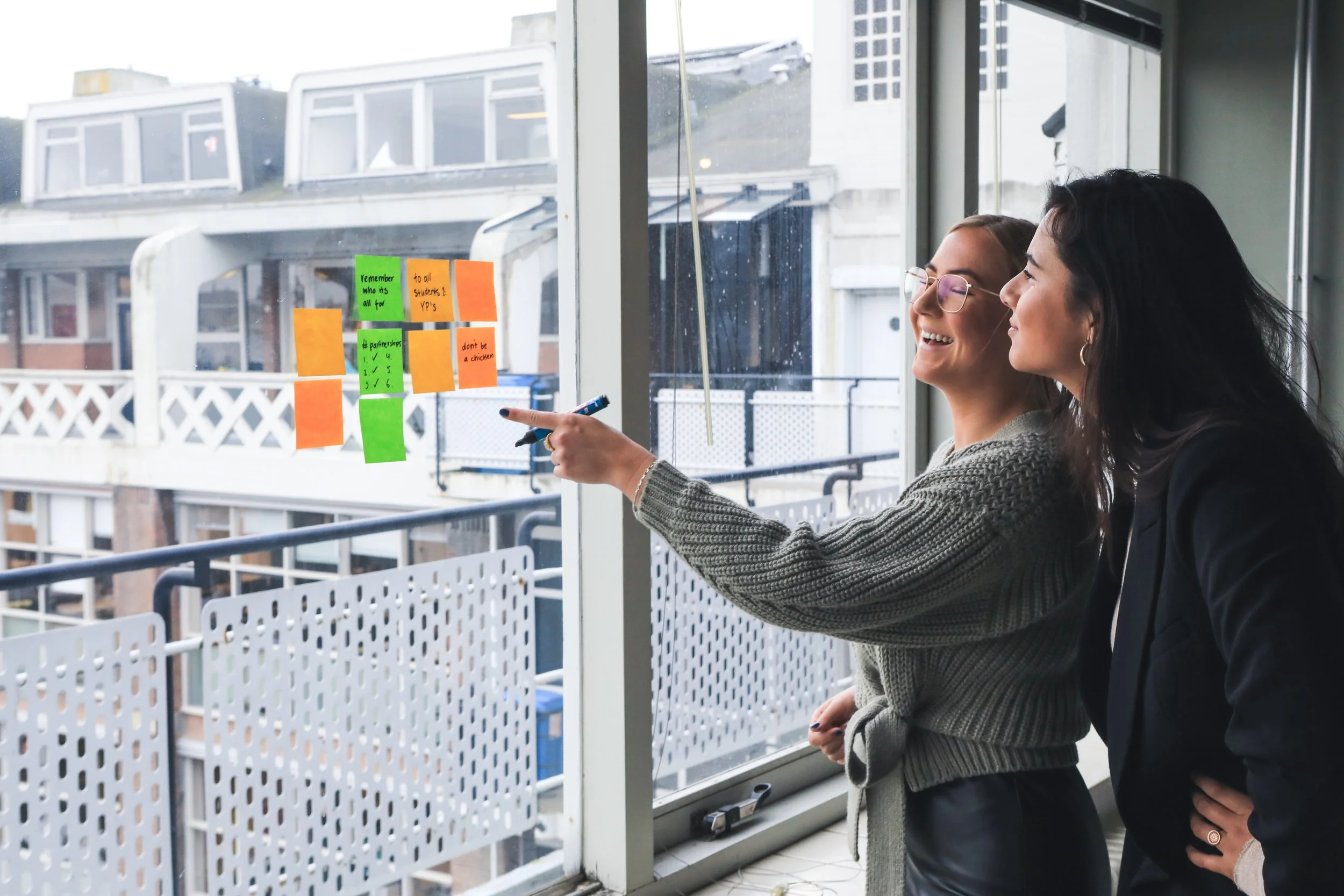 Two women standing by a large window with colorful sticky notes on the glass, smiling and talking.