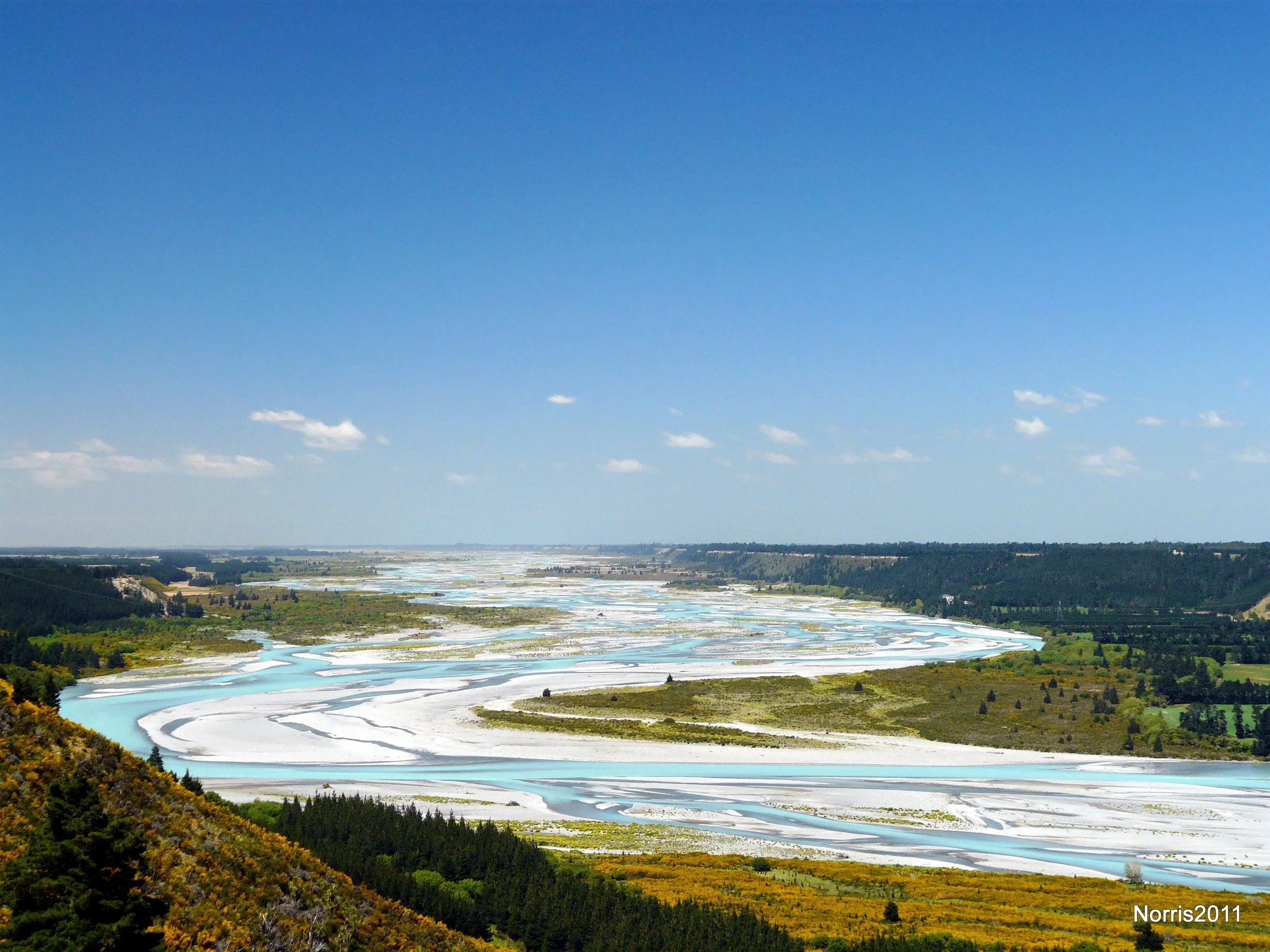 View of a river winding through lush green landscape with patches of yellow and white sand, under a blue sky with scattered clouds.
