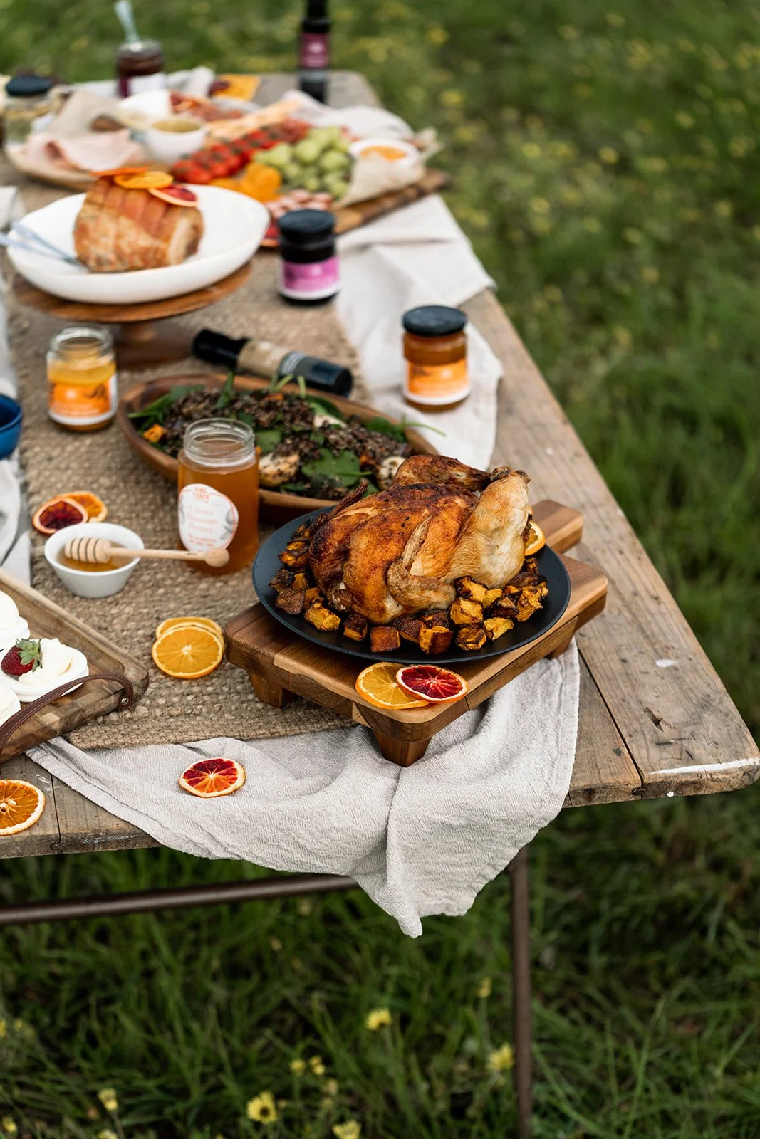 Kingfisher Citrus preserves and condiments displayed on a rustic table with a white tablecloth, outside. On the table is a roast chicken, salads, glazed ham and more.