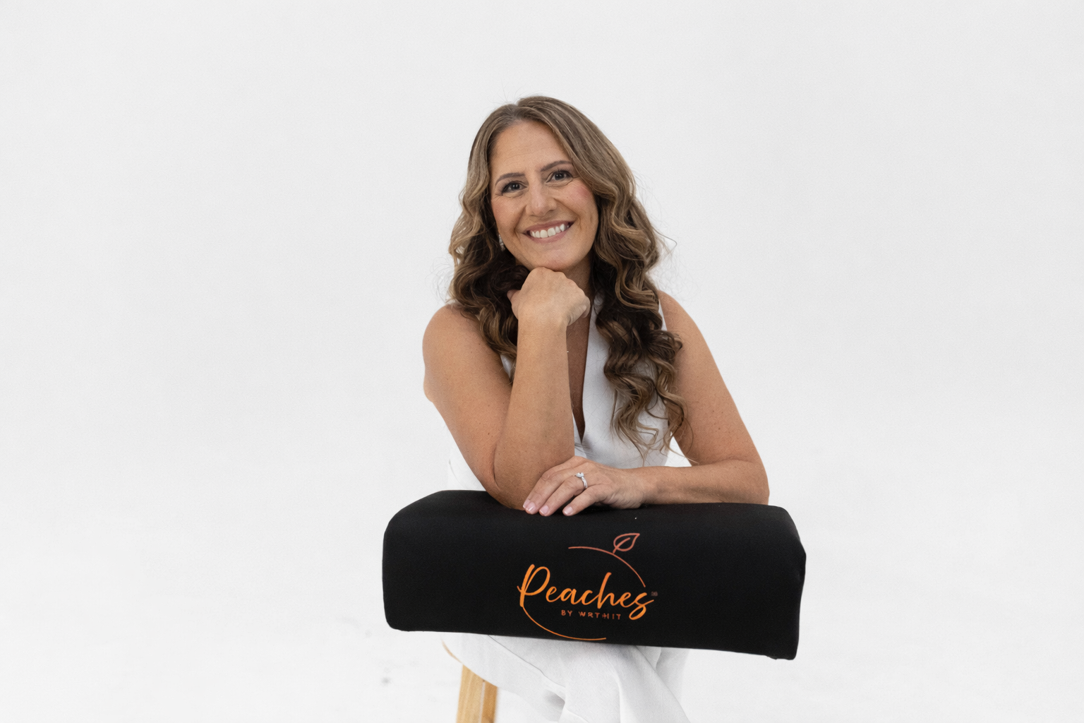 A smiling woman with curly brown hair sitting on a stool holding a bbl pillow with a logo that reads "Peaches by Writht" in orange font, against a plain white background.