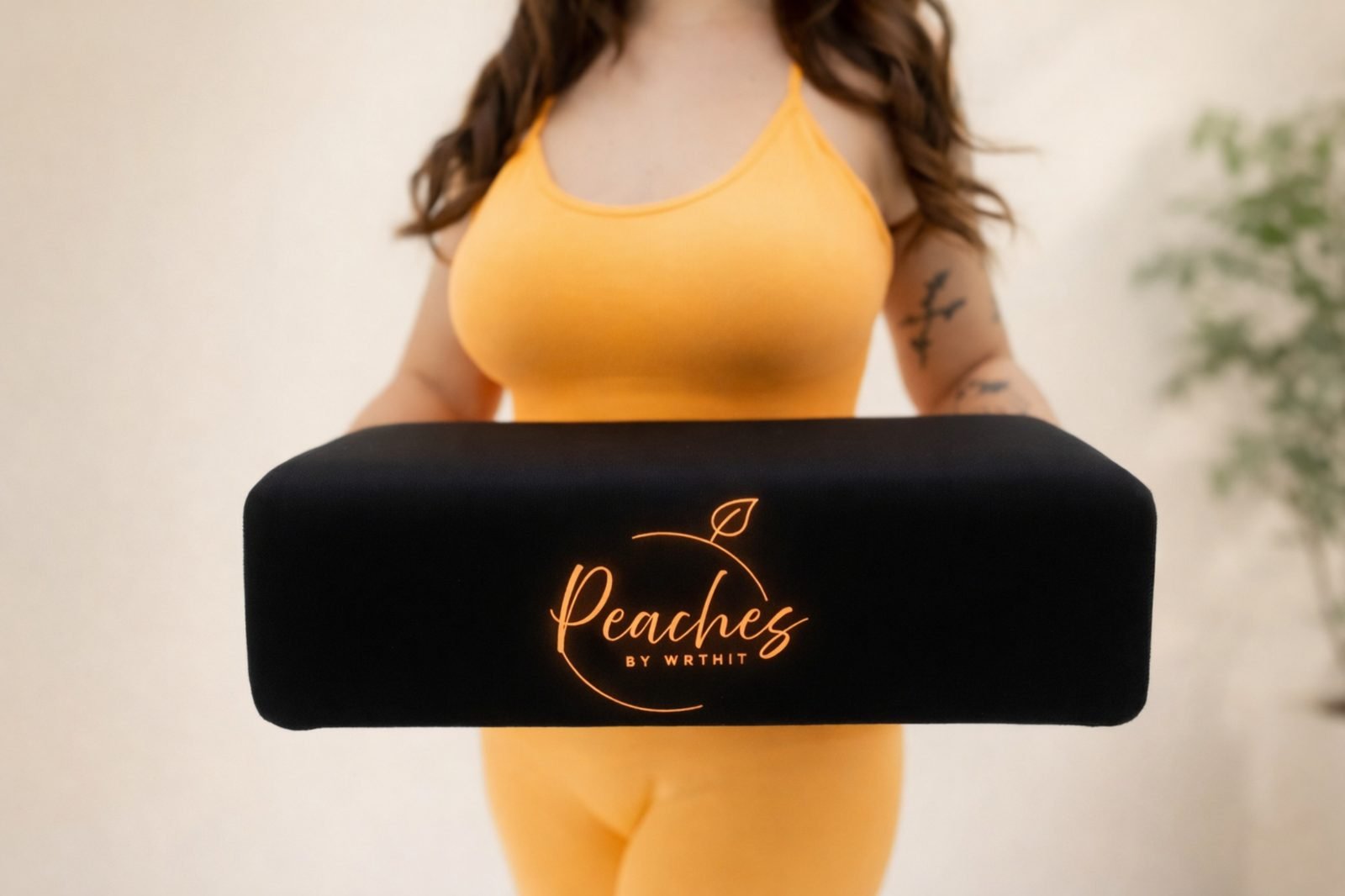 Woman in orange workout clothes holding black BBL pillow called The Bellini from Peaches by Wrthit