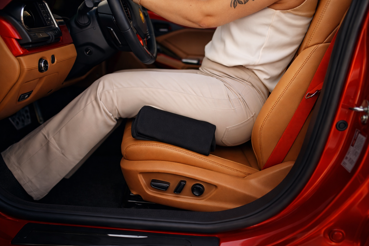 Interior of a car showing a person sitting in the driver's seat with tan leather upholstery, beige pants, and a white shirt, sitting on a bbl pillow