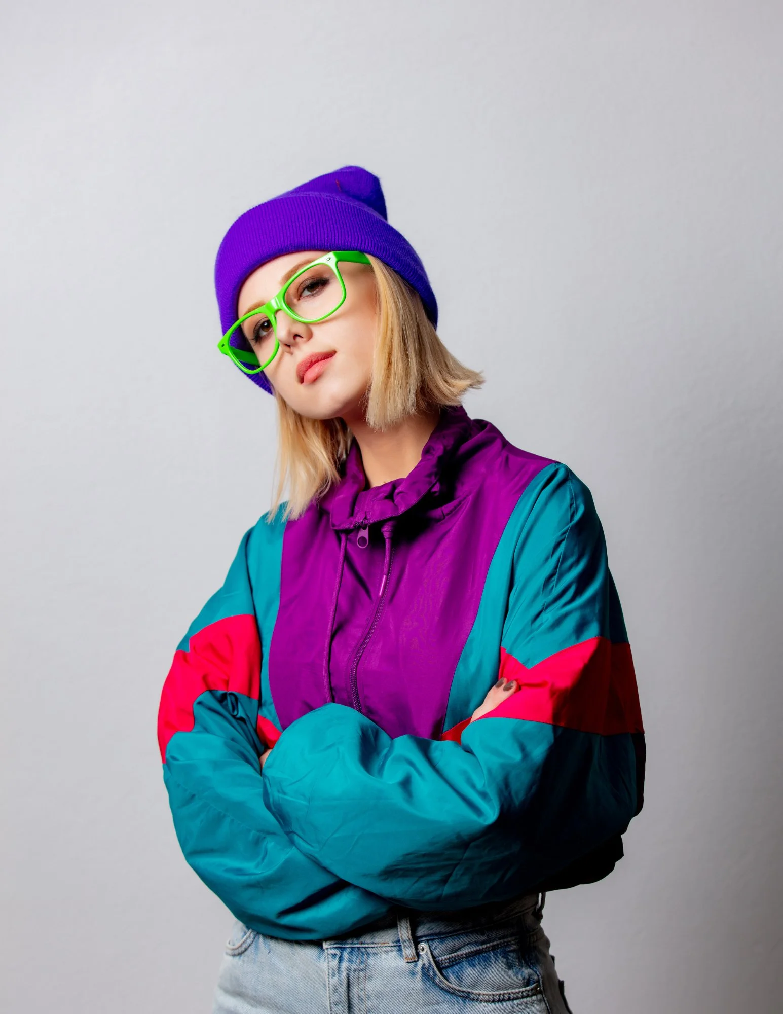 A young woman with blonde hair wearing a purple beanie, green glasses, a colorful windbreaker, and light blue jeans, standing against a plain gray background.