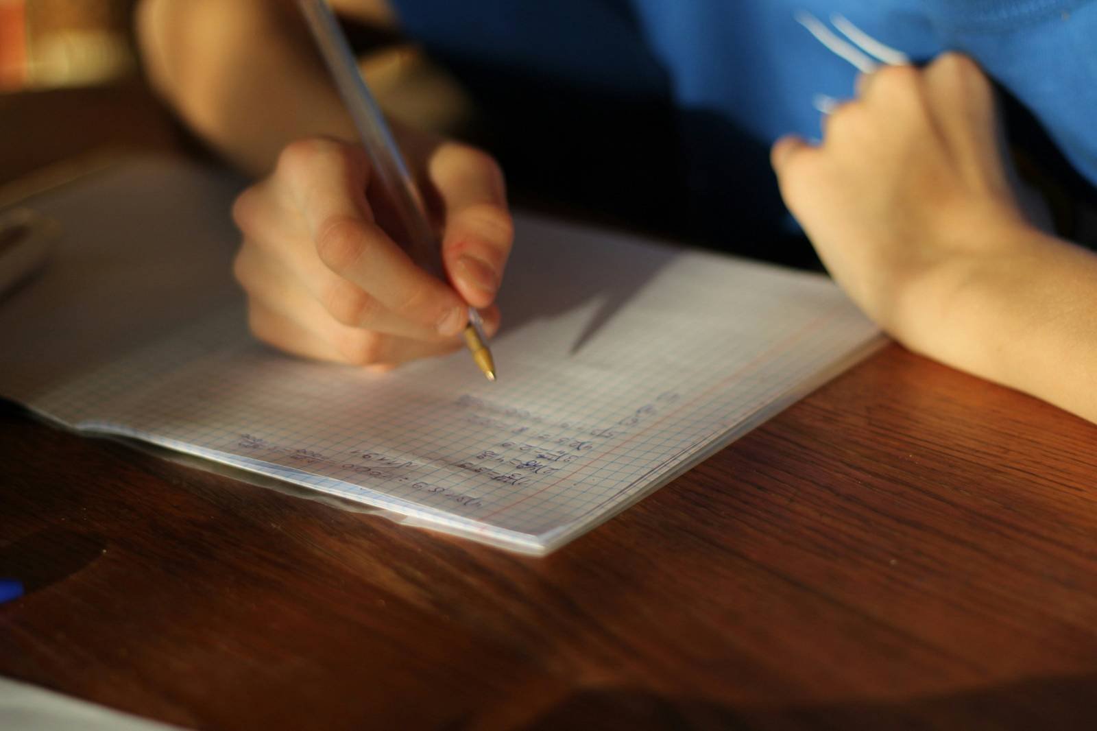 Close-up of a person's hand writing in a notebook on a wooden table, using a gold-colored pen on graph paper.
