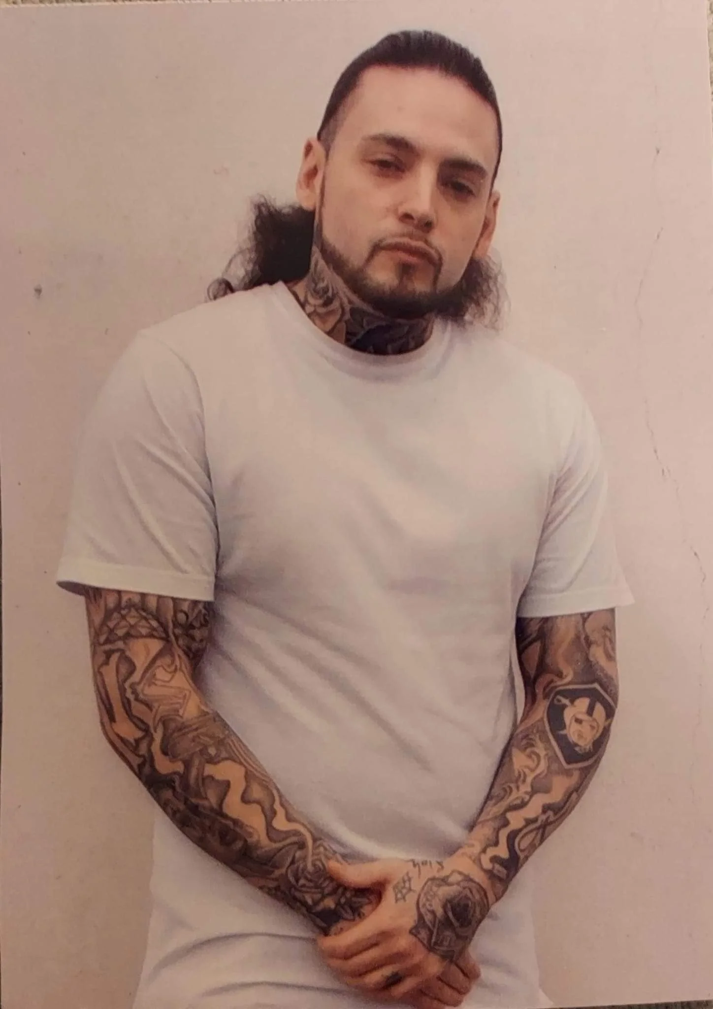 A man with tattoos on his arms and neck, wearing a plain white T-shirt, standing against a light-colored wall.
