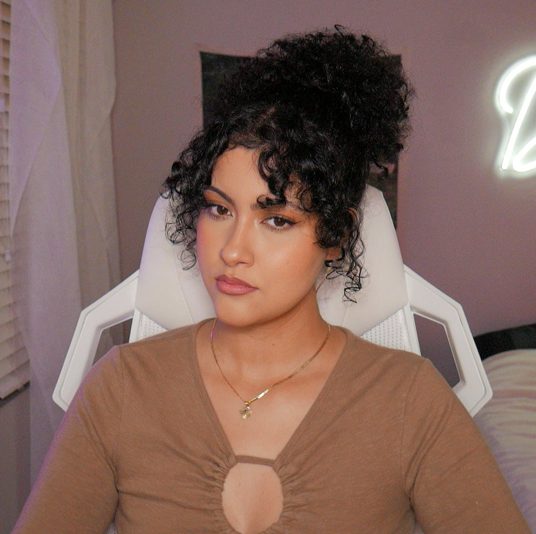 A woman with curly dark hair sitting on a white chair with a high backrest, in a room with soft lighting, wearing a brown top with a keyhole cutout, and gold jewelry.