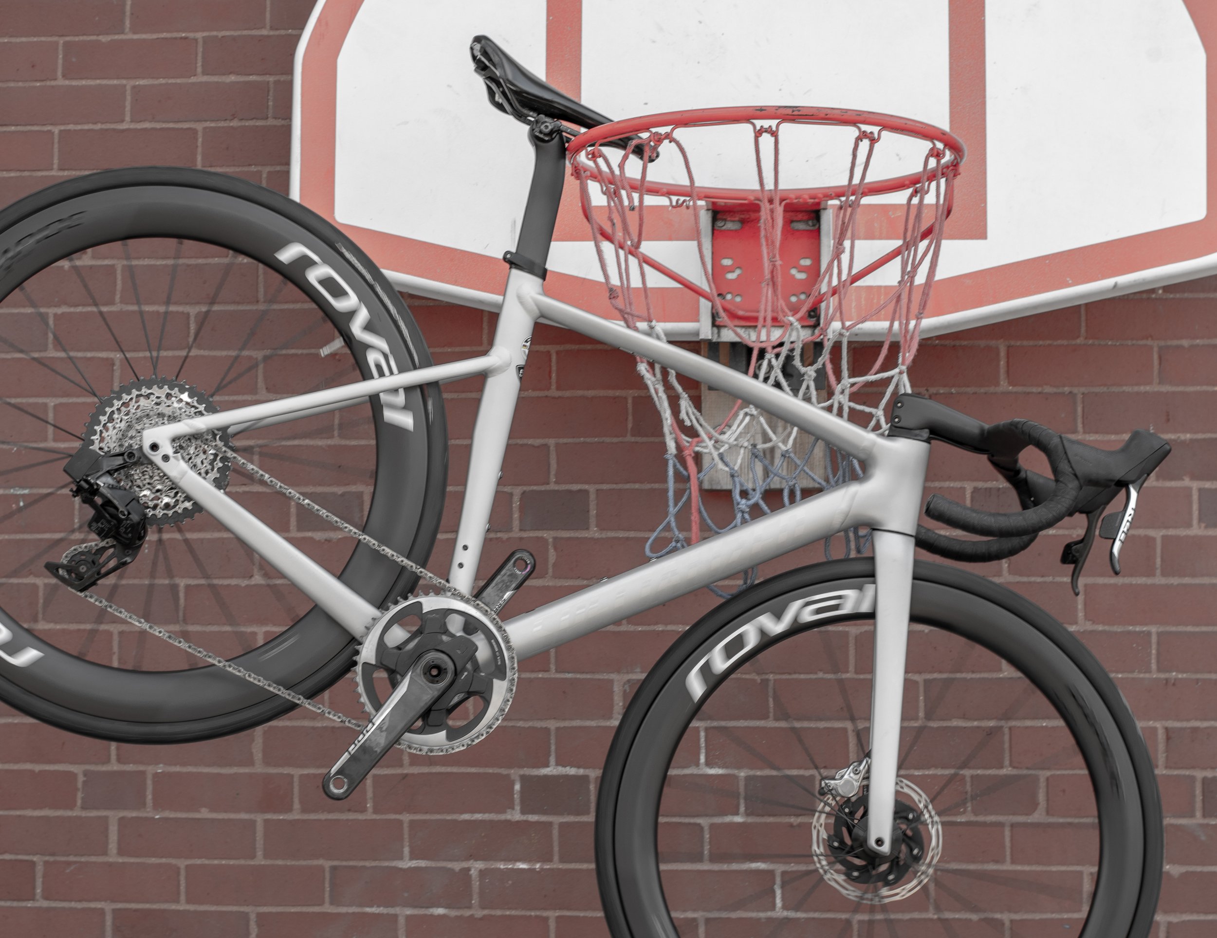 A silver road bicycle with black tires and a red basketball hoop attached to a brick wall in the background.