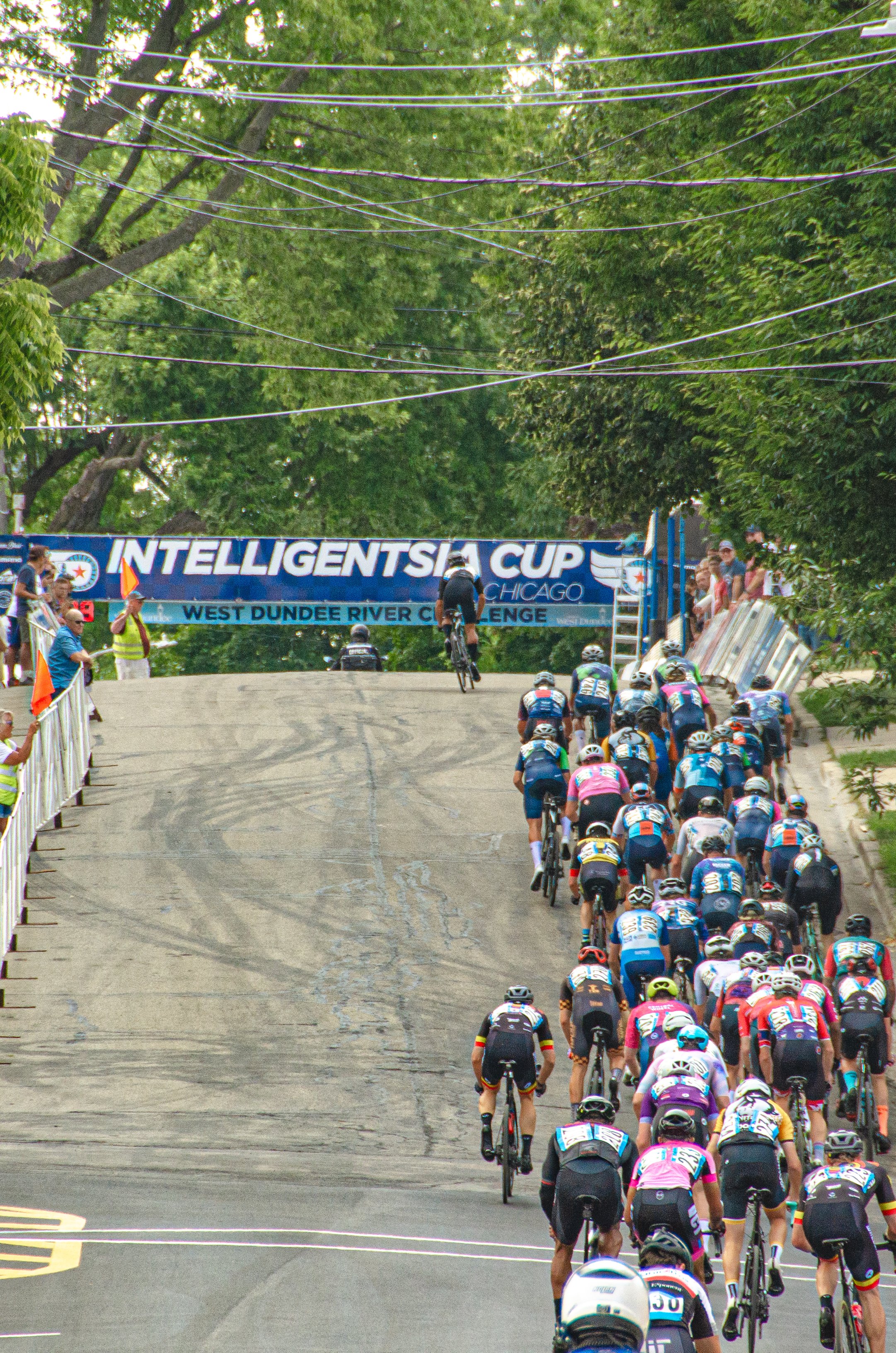 Cyclists competing in the Intelligentisa Cup bike race on a hilly road, with spectators cheering on the side and banner overhead, surrounded by trees.