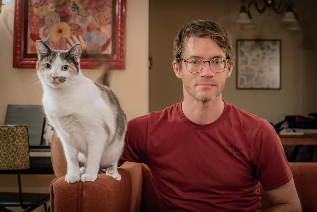 A man with glasses and a red shirt sitting on a couch with a white and gray tabby cat standing on the armrest beside him in a cozy living room.