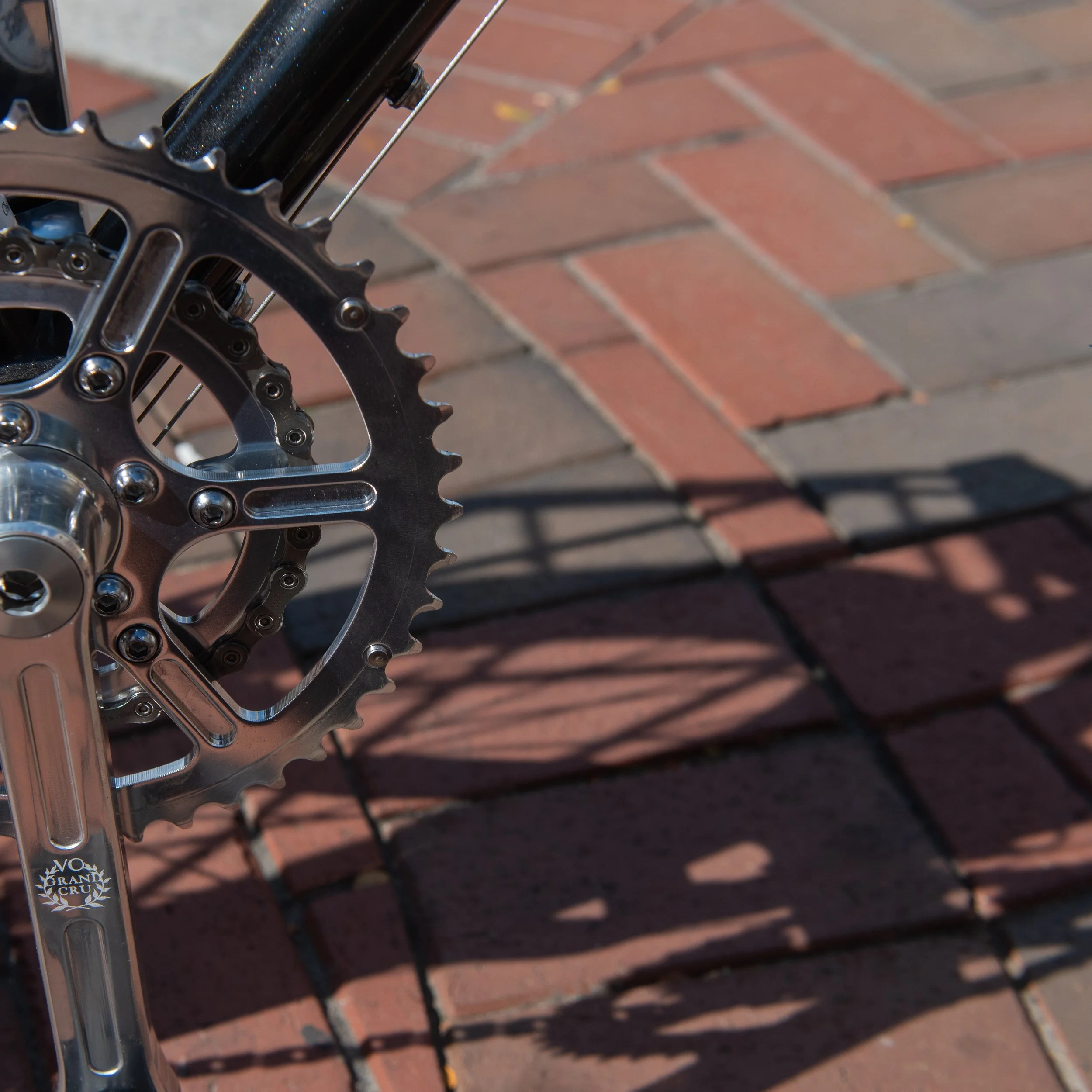 Close-up of a bicycle crankset and chainring with sunlight casting shadows on a brick pavement.