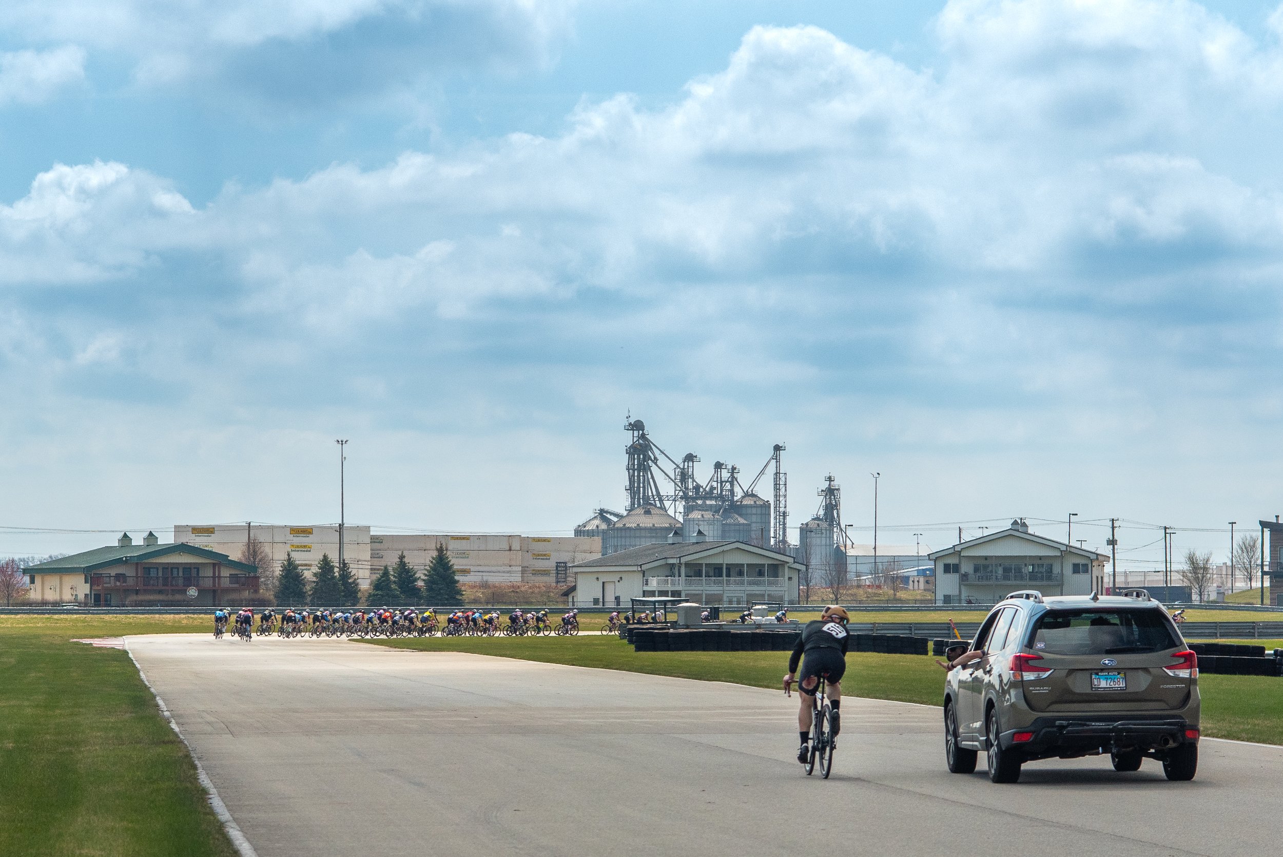 A cyclist riding on a race track with a backup vehicle behind them, with a large group of cyclists in the distance, and industrial structures or silos in the background under a partly cloudy sky.