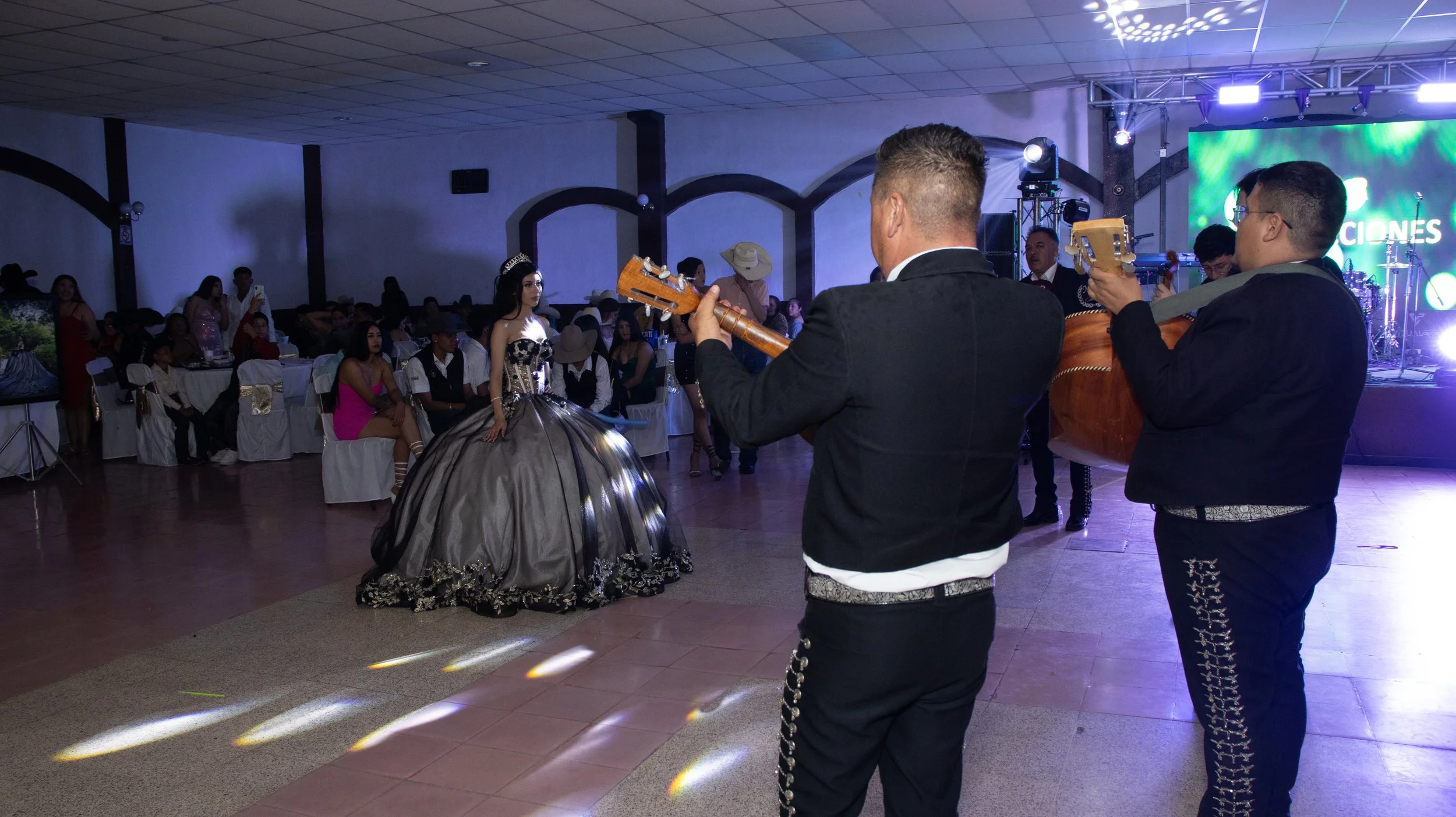 Buscamos un encuadre narrativo: colocamos al mariachi en primer plano para conducir la mirada hacia la quinceañera en luz puntual. La atmósfera del salón, con haces de iluminación y público al fondo, refuerza la emoción de la presentación.