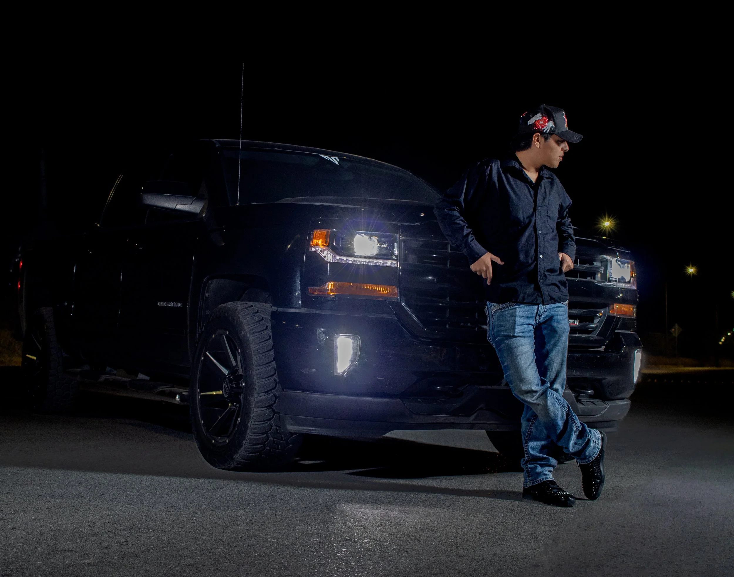 El protagonista irradia fuerza y estilo, apoyado casualmente en su Silverado negra. Su postura relajada y mirada lateral proyectan confianza y dominio, elementos esenciales de nuestra estética urbana nocturna.