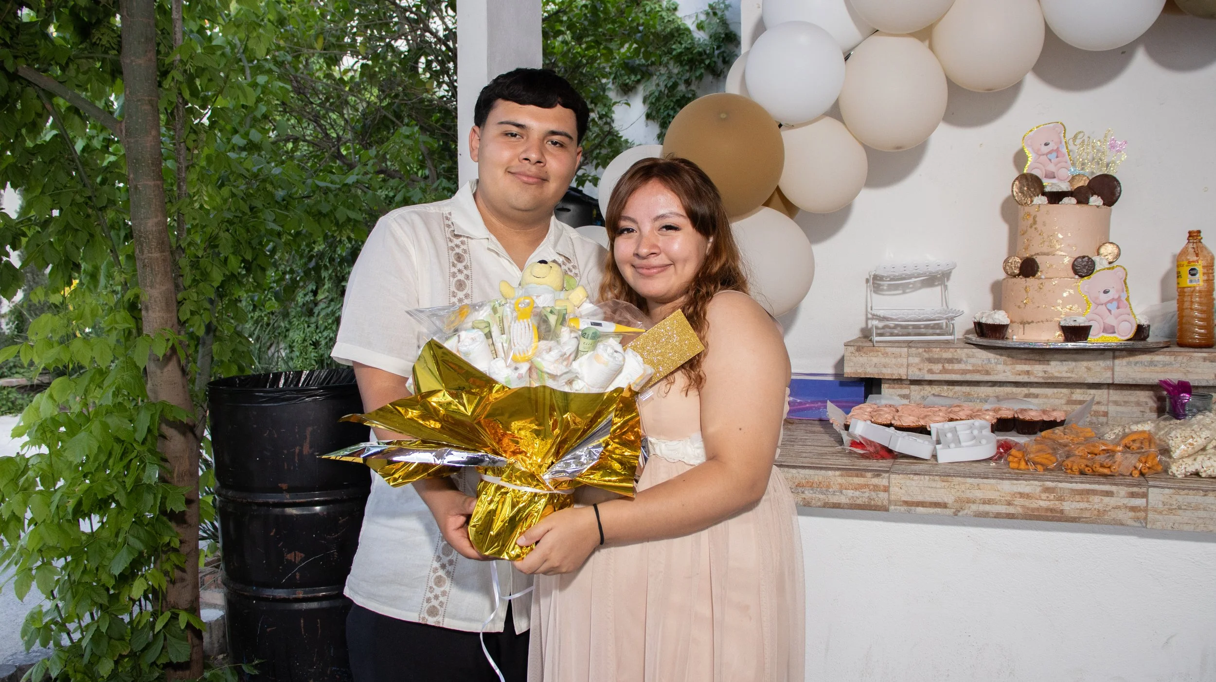 Optamos por una iluminación controlada que preserve el ambiente del salón y, a la vez, defina texturas: el dorado del ramo, el glaseado del pastel y los globos del fondo. Un retrato cálido que celebra la generosidad de los invitados y el cariño de la