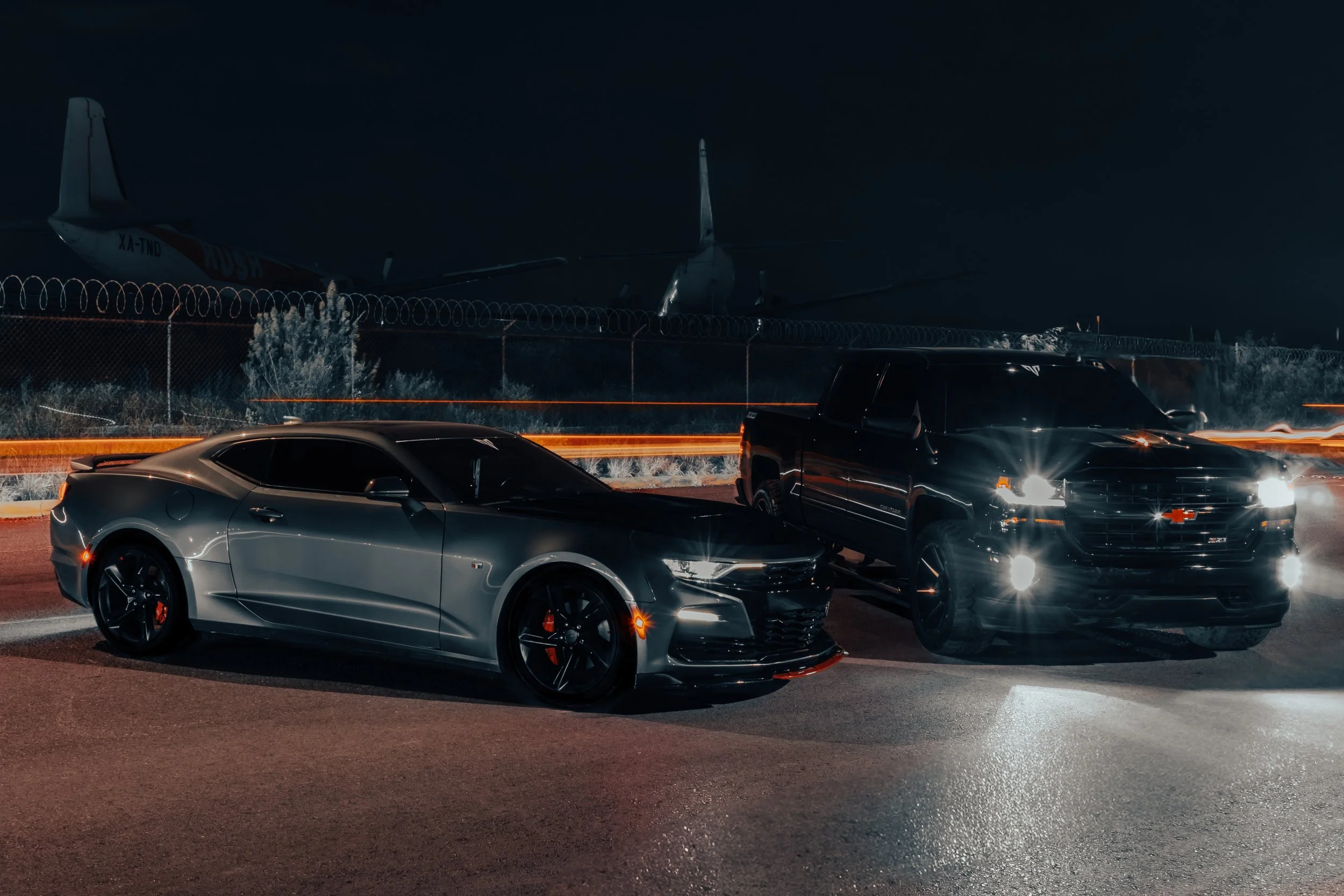 Toma cinematográfica que combina la potencia del Camaro gris y la Silverado negra con un fondo industrial. Reflejos metálicos, luces cálidas en movimiento y un rastro de luz simbolizan velocidad.