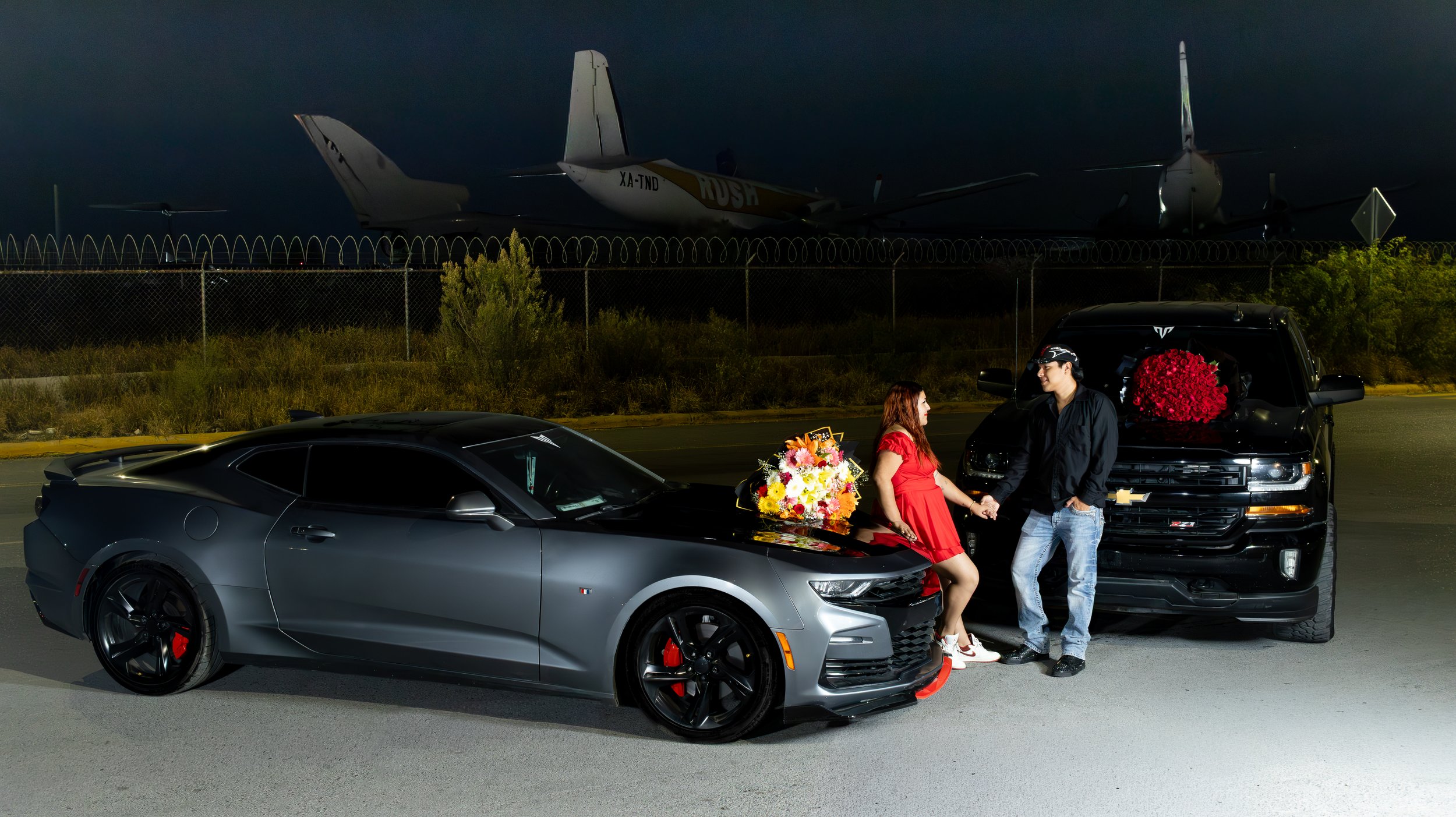 La pareja, vestida en tonos complementarios, enmarcada por los vehículos, luces y detalles florales. Dualidad entre fuerza automotriz y conexión humana.