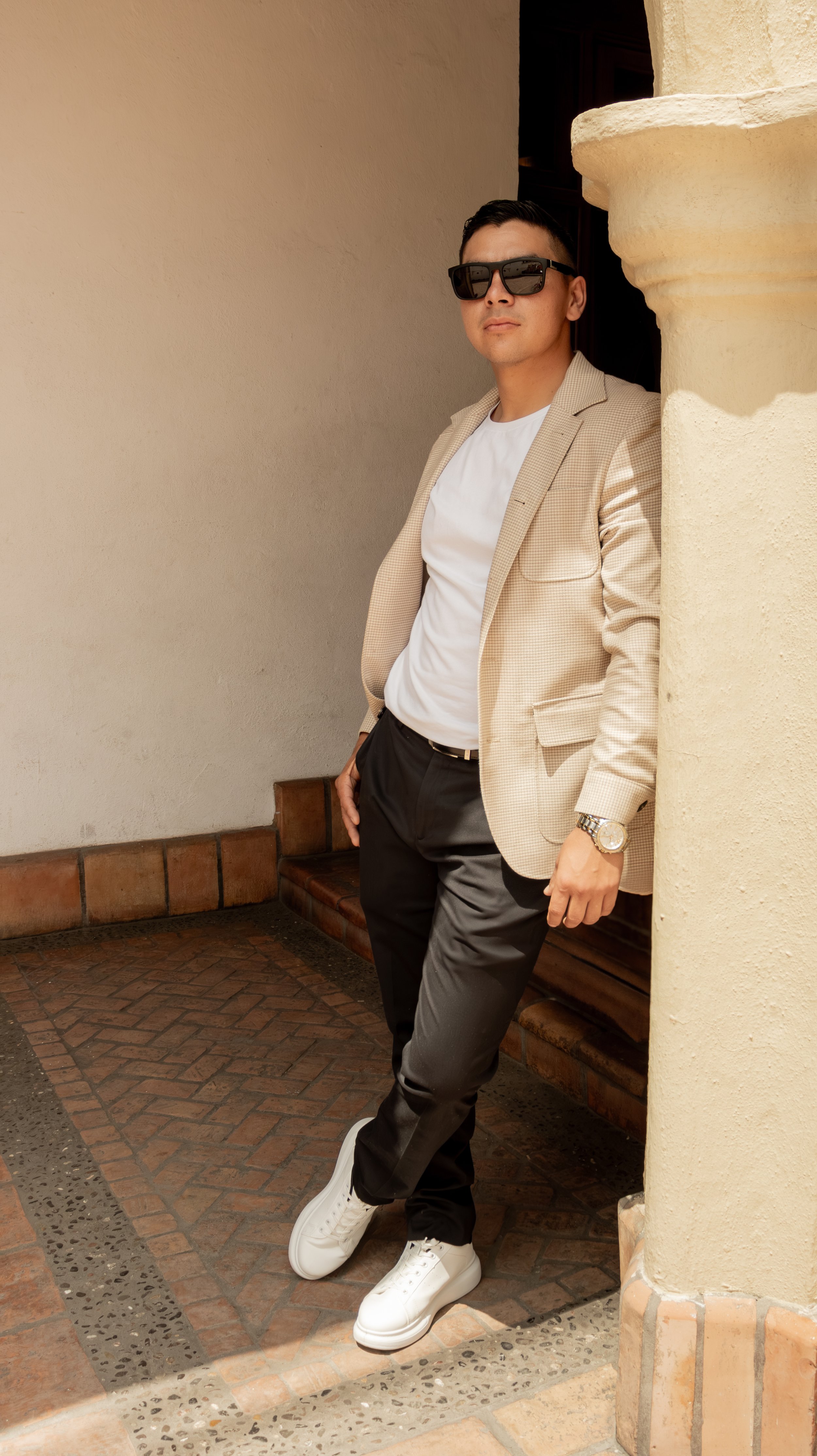 Columna como marco vertical, piernas cruzadas y peso al muro para una pose relajada. La luz de mediodía se difumina sobre el saco y zapatillas blancas para un contraste moderno.
