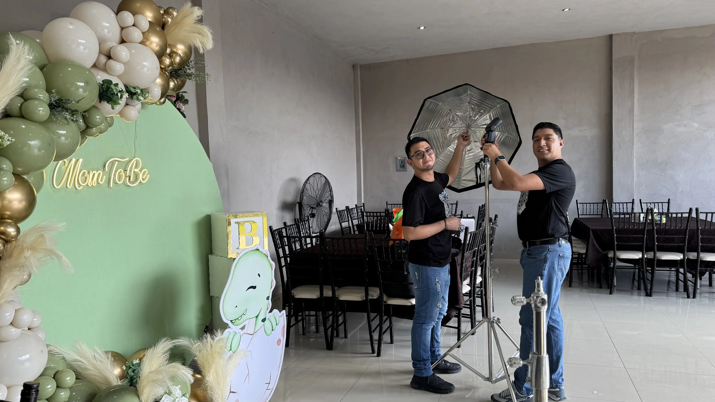 Dos hombres ajustando una iluminación en una sala decorada para un evento con un cartel que dice 'Mom To Be' y decoraciones en tonos verde, blanco y dorado.