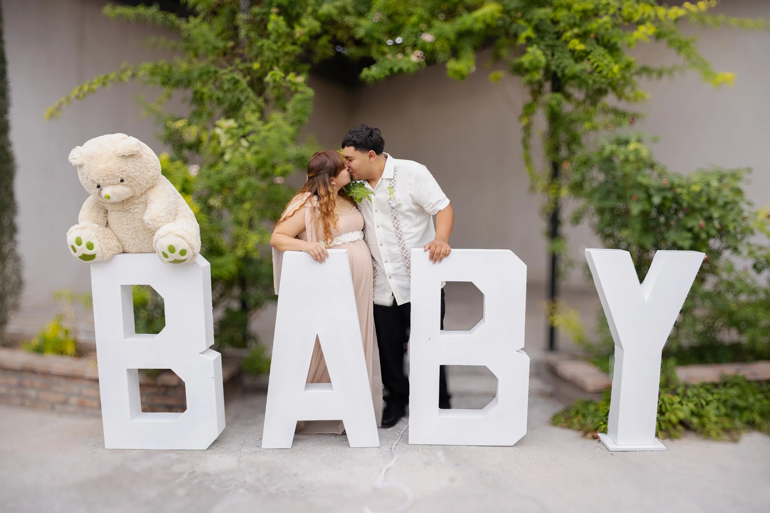 Buscamos un encuadre lúdico y limpio, usando las letras gigantes como líneas guía para dirigir la atención al beso central. Desenfocamos el fondo para dar protagonismo a la pareja y mantener el ambiente tierno con el peluche como acento.