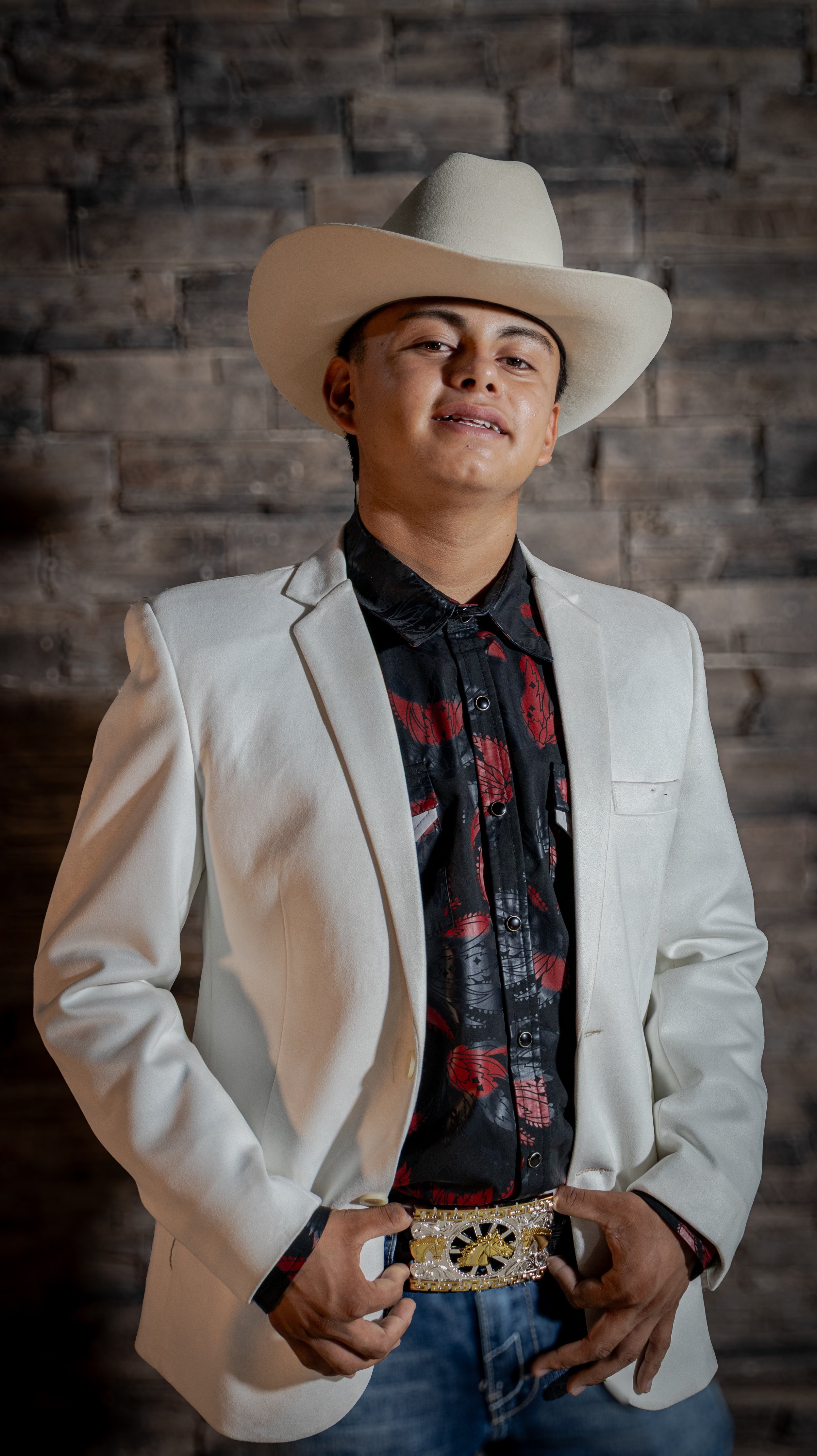 Iluminación dirigida y fondo de piedra para texturas limpias. Posamos al festejado con saco blanco y sombrero, destacando la hebilla y el gesto seguro para un retrato clásico y elegante.