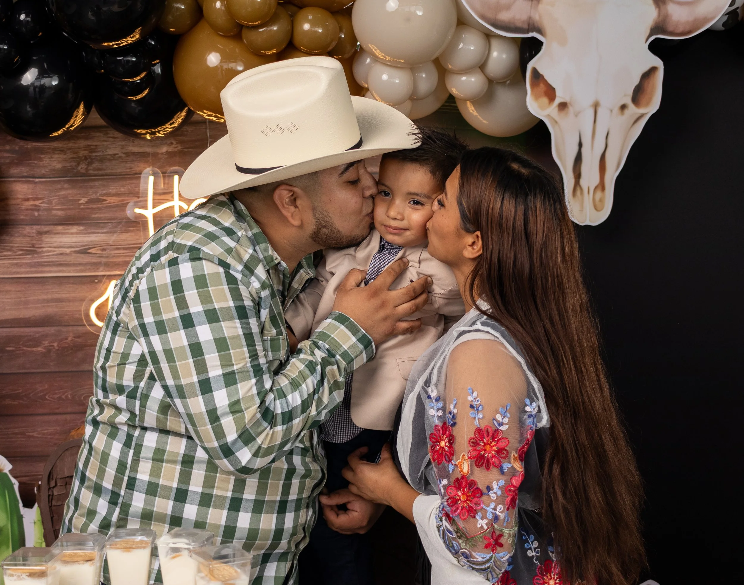 Enmarcamos el abrazo y los besos de papá y mamá con el pequeño al centro, usando profundidad reducida para centrar la atención en el gesto de cariño.