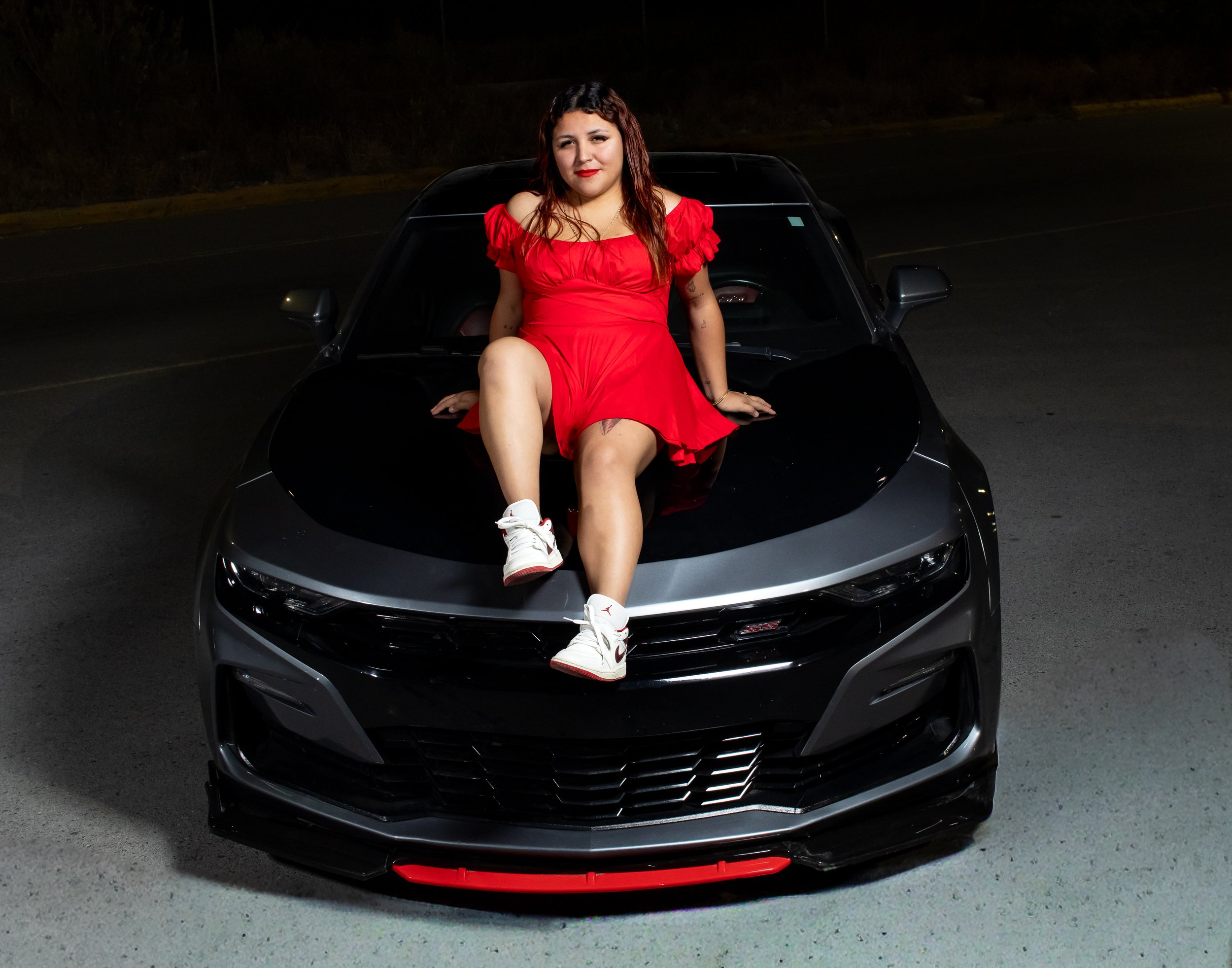 La modelo sobre el Camaro, mezclando feminidad, fuerza y estilo. Luz puntual acentúa el contraste del vestido rojo con el acabado metálico, transmitiendo poder y elegancia.