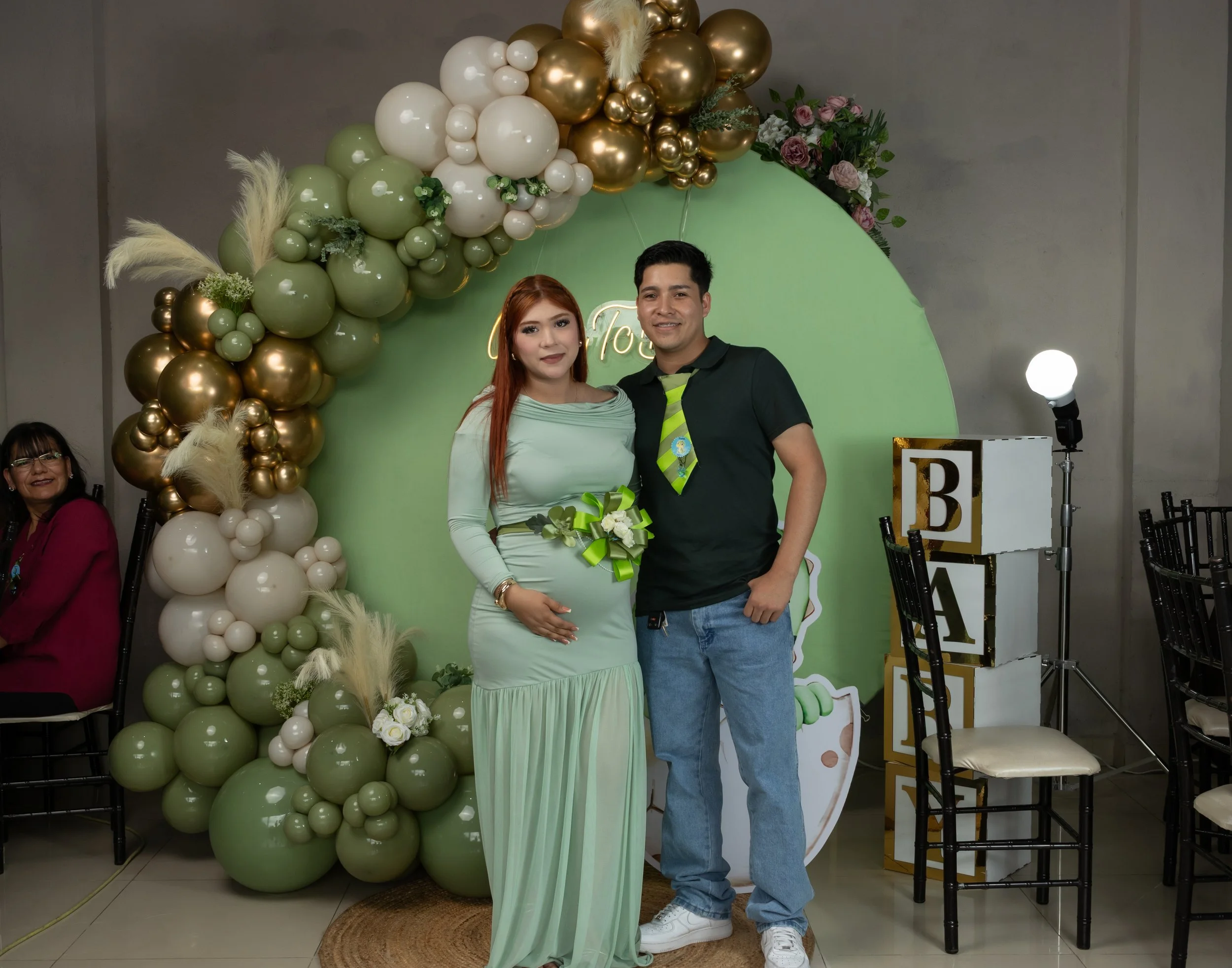 Creamos un retrato limpio y elegante bajo el arco de globos verde/oro. Luces suaves, composición centrada y manos sobre el vientre para contar, en una sola imagen, la historia que empieza.