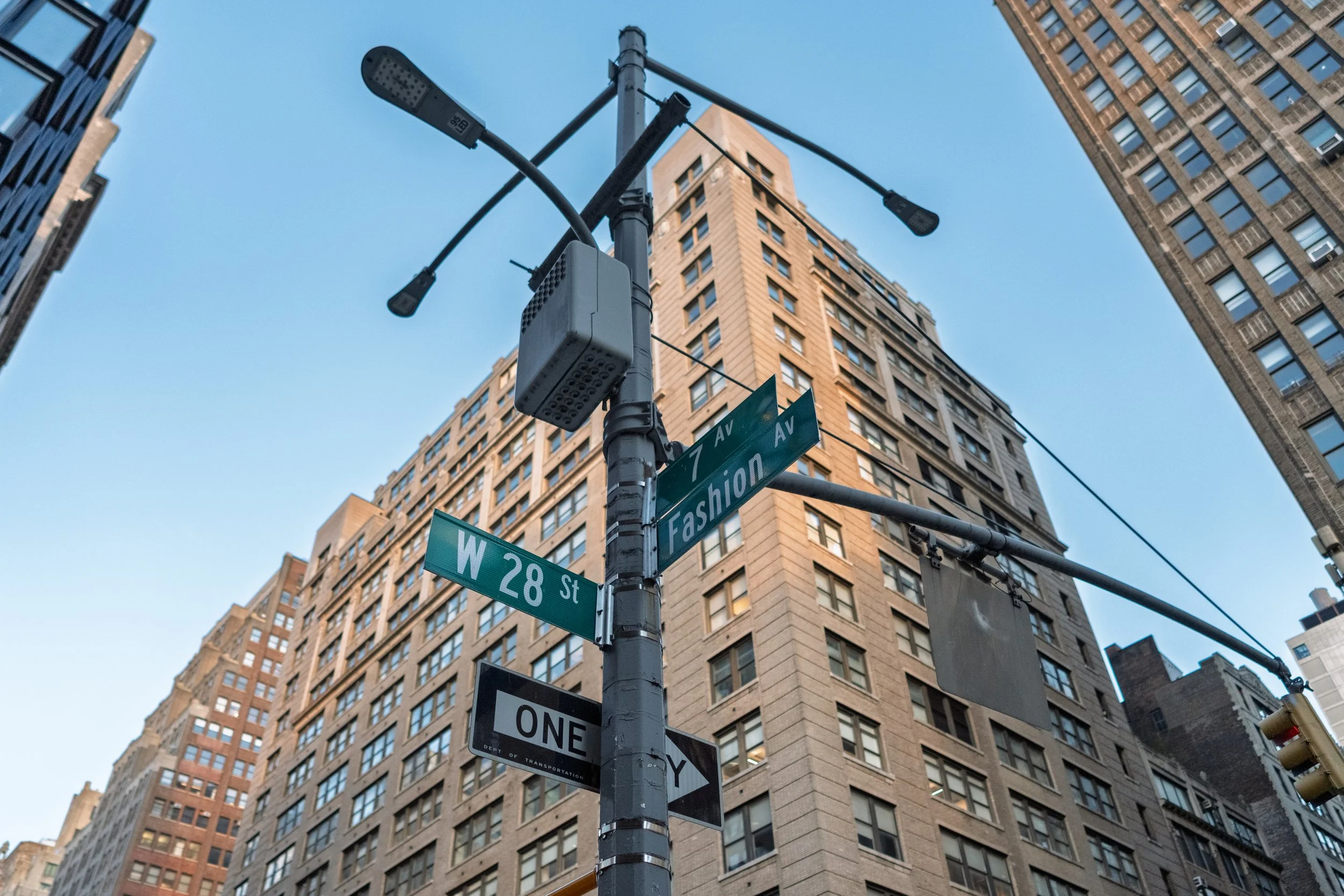 Street corner in New York City, with traffic light and street signs for W 28th Street and Fashion Avenue, surrounded by tall buildings with windows.