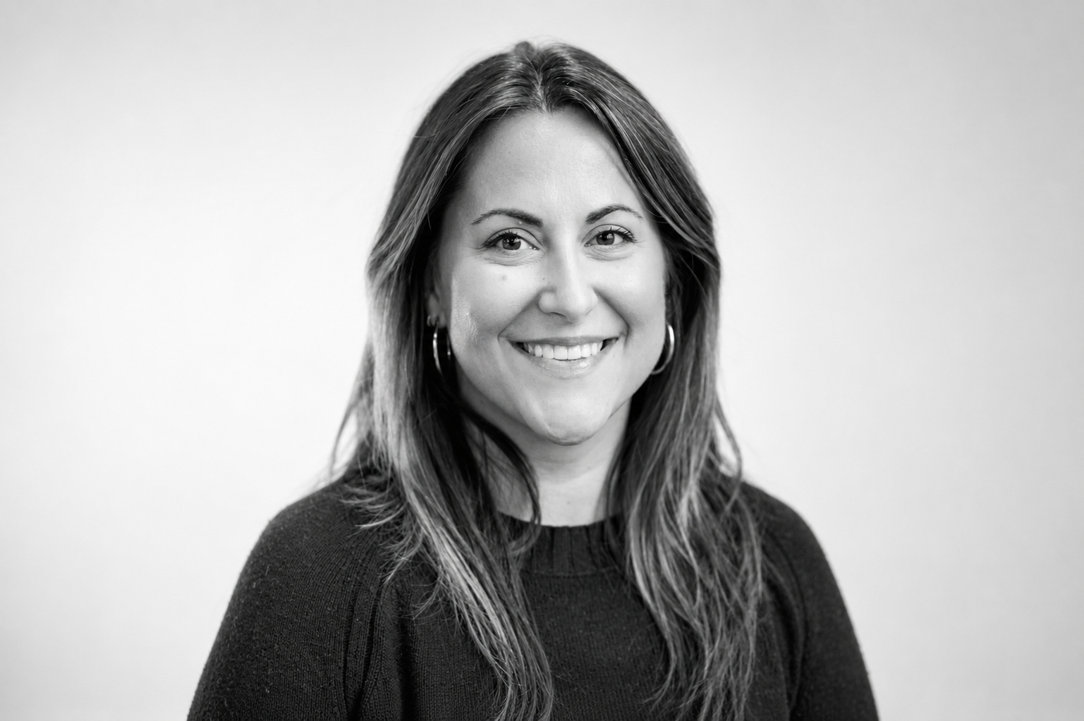 Black and white portrait of a smiling woman with long hair, wearing a necklace and a dark top.
