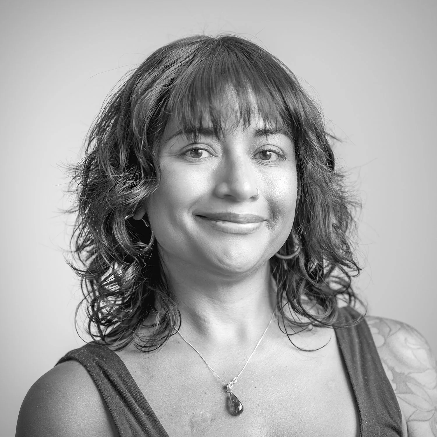 Black and white portrait of a woman with short dark hair, wearing hoop earrings, sleeveless top, and displaying a tattoo on her left arm against a plain background.