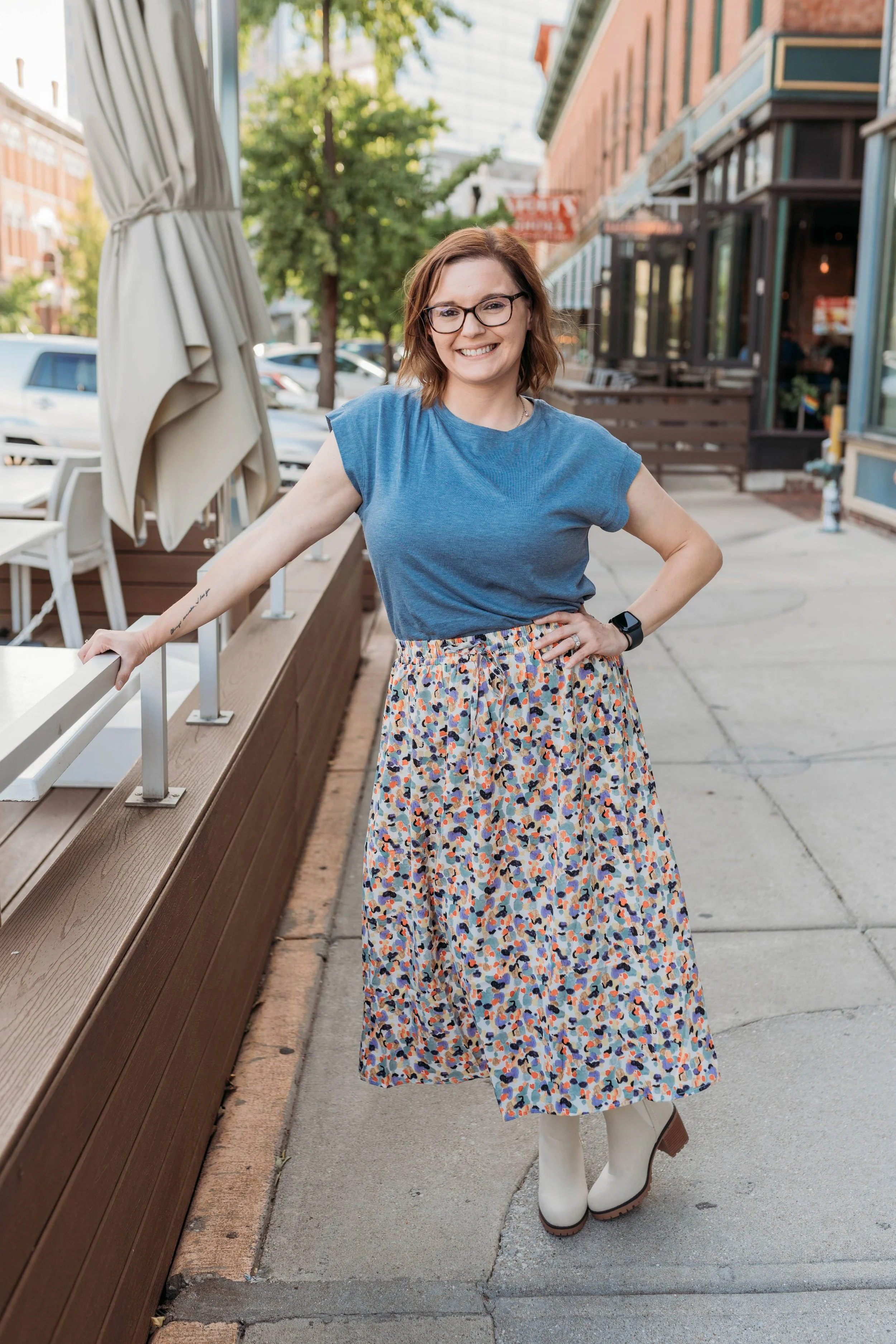 A smiling woman with glasses and shoulder-length brown hair posing outdoors on a sidewalk, wearing a blue t-shirt, a colorful floral skirt, and white boots, with a wooden bench and patio umbrellas behind her.