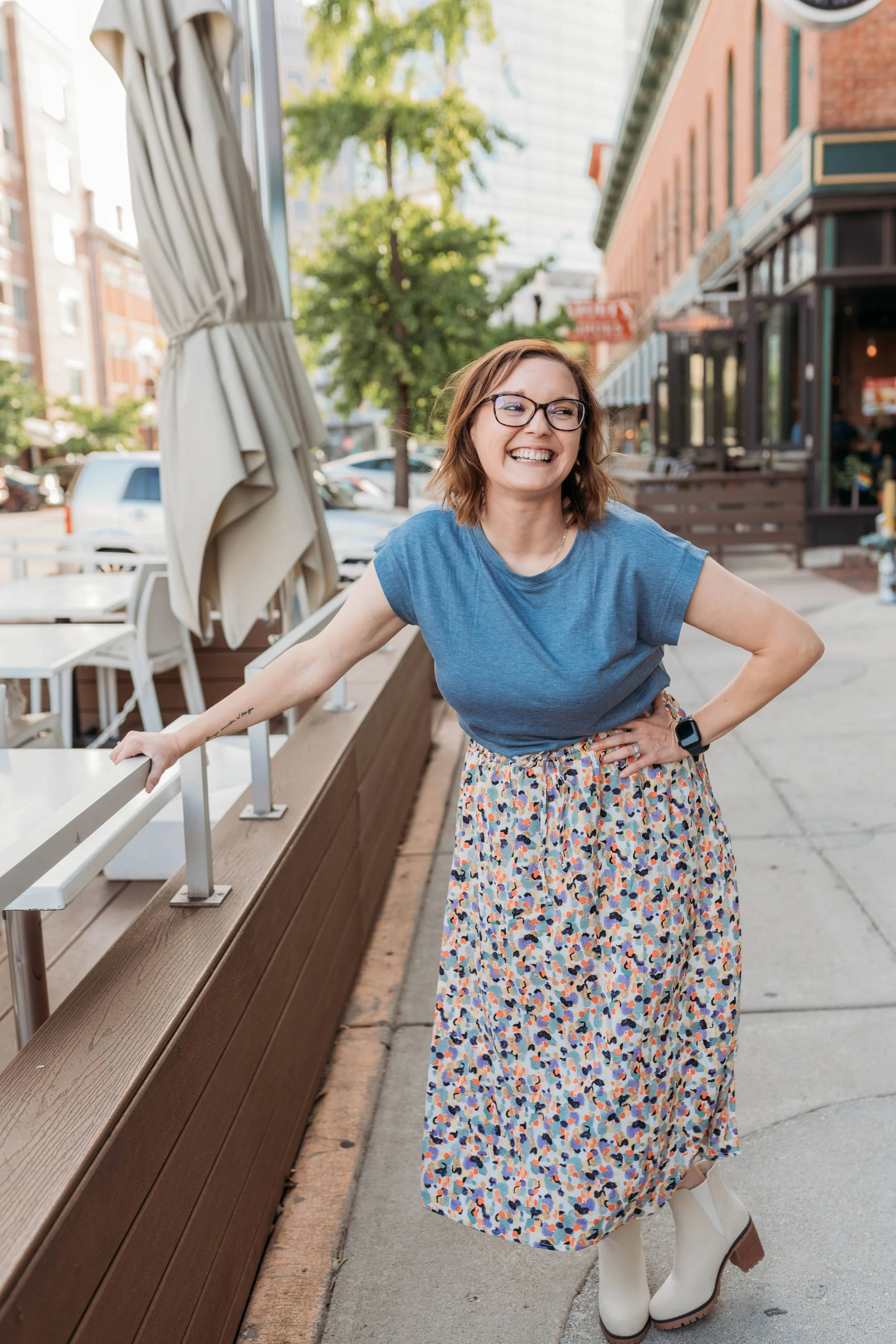 A woman with glasses and wavy brown hair smiling brightly, wearing a blue T-shirt, a colorful patterned maxi skirt, white ankle boots, and a smartwatch, standing outdoors on a city sidewalk near a brown wooden bench and a beige umbrella. Trees, parked cars, and storefronts are visible in the background.