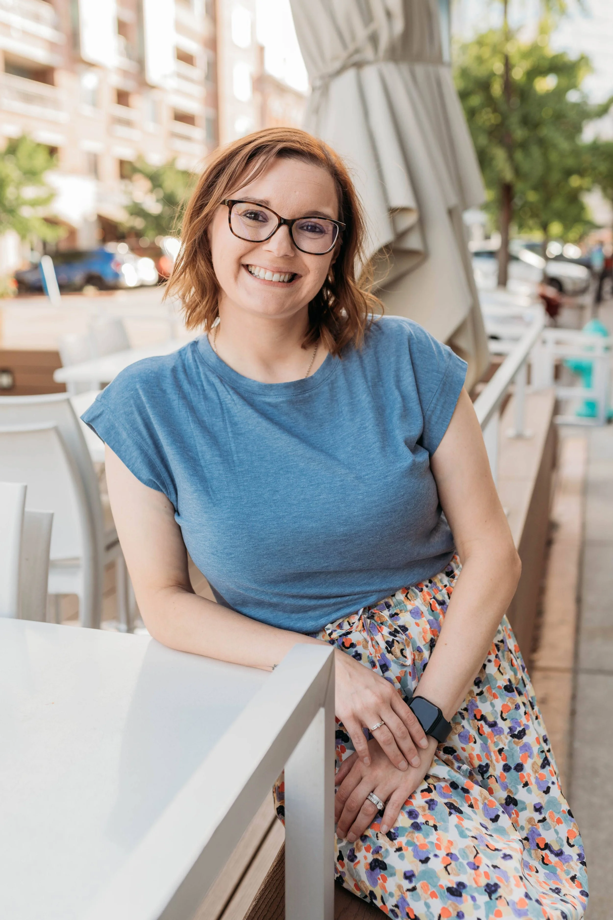A woman with glasses smiling, sitting outdoors at a white table, wearing a blue top and a colorful patterned skirt, near a beige umbrella with buildings and trees in the background.