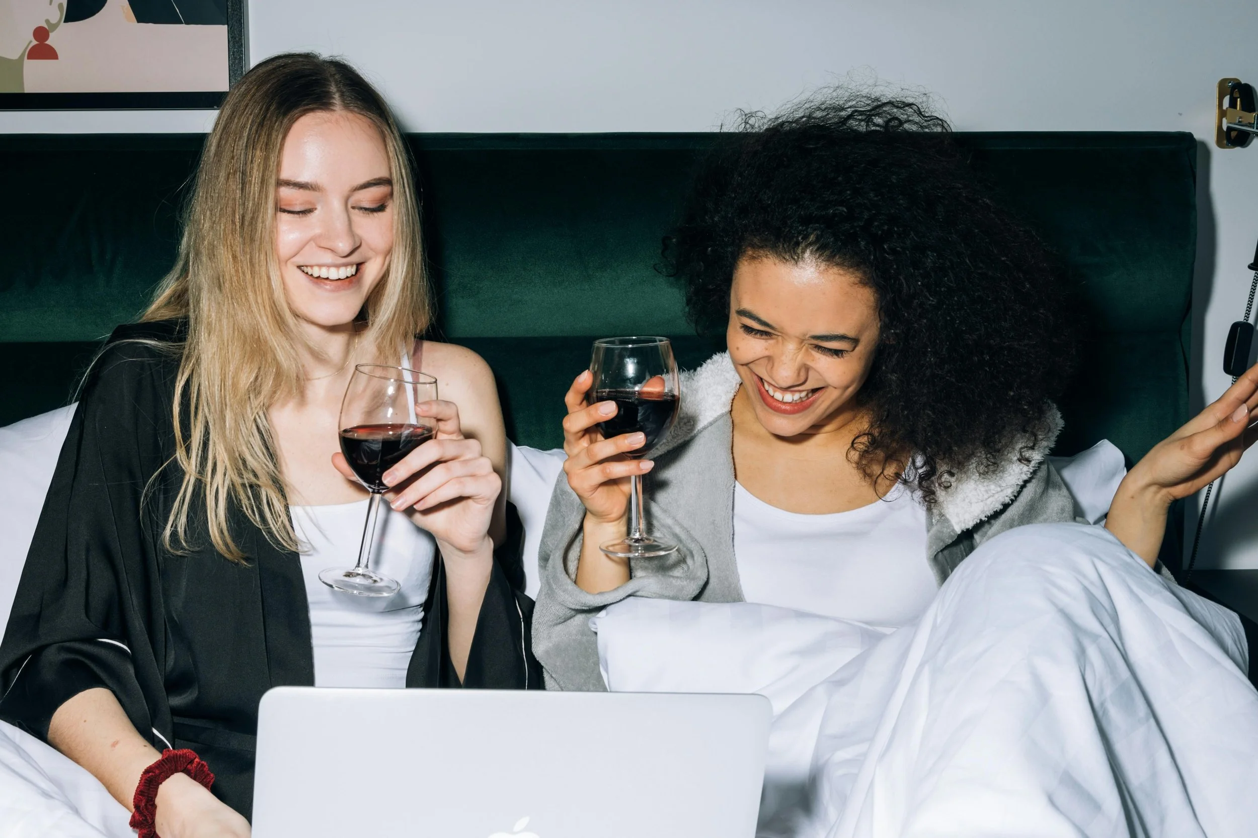 Two women sitting on a bed, laughing and holding glasses of red wine, with a laptop in front of them, in a bedroom setting.