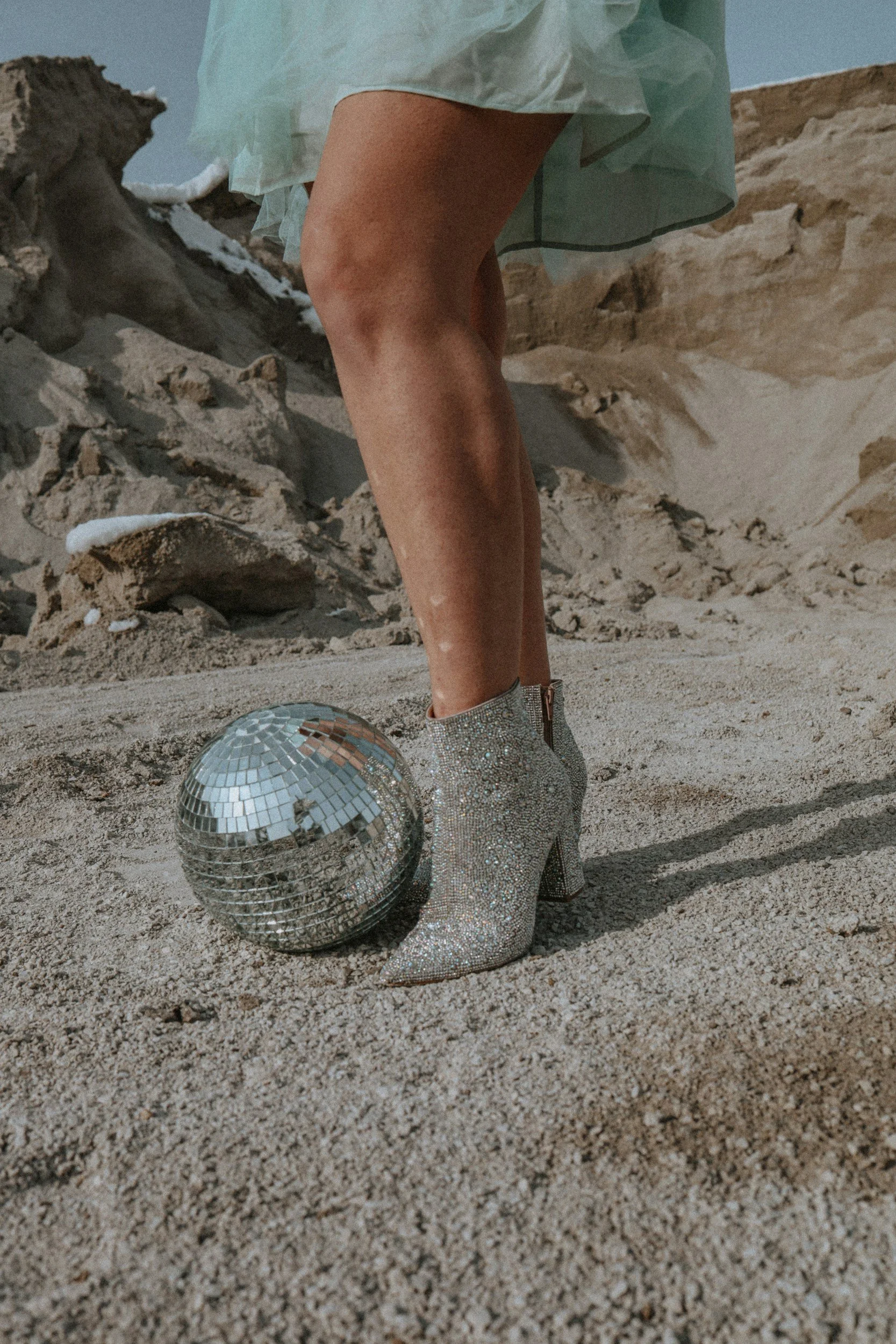 Close-up of a person's legs and feet wearing sparkling silver high-heeled boots on sandy ground, with a disco ball nearby. The person is dressed in a light-colored skirt, with rocky formations in the background.