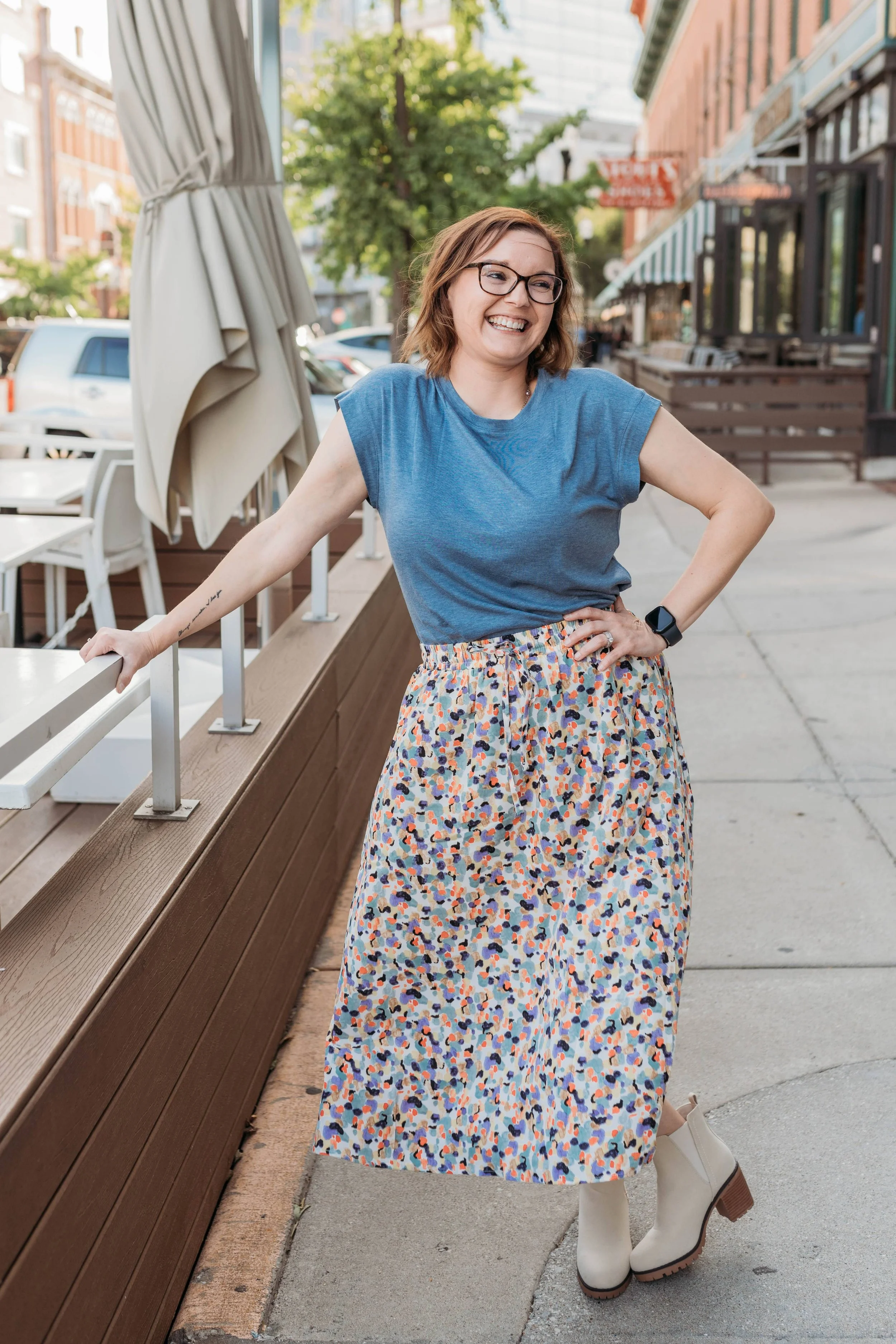 A woman with shoulder-length brown hair, glasses, and a bright smile stands outside on a city sidewalk. She is wearing a blue sleeveless top, a colorful patterned skirt, and white boots with a thick heel. She has her left arm on her hip and her right arm resting on a wooden ledge. In the background, there are storefronts, parked cars, and greenery.