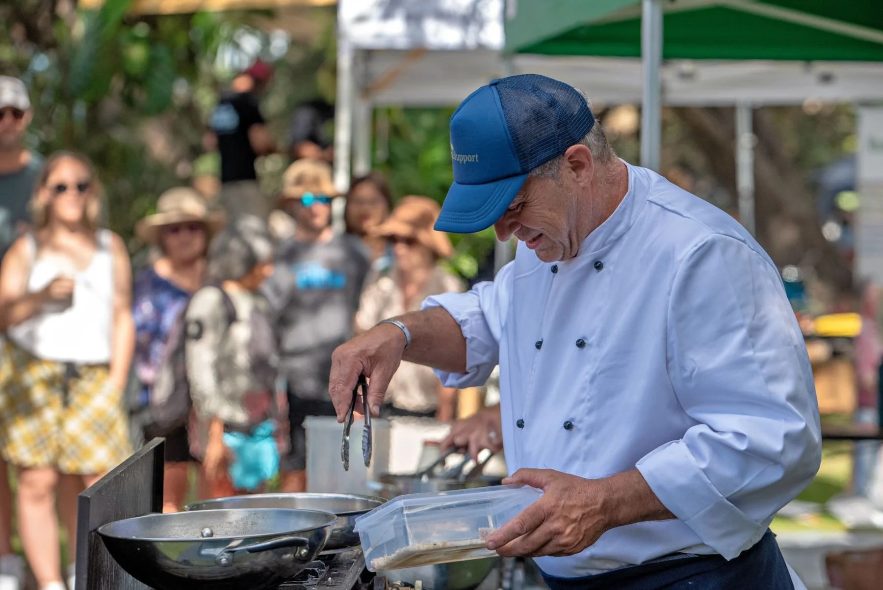 Chef in a white chef's coat and blue cap cooking outdoors at a food event, with a crowd of people watching in the background.