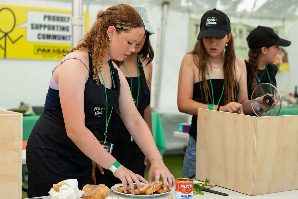 Young women participating in mystery box cooking challenge, preparing sandwiches at a table. They are wearing black aprons and green wristbands, with some also wearing caps. One woman appears focused on making a sandwich with bread and spread.