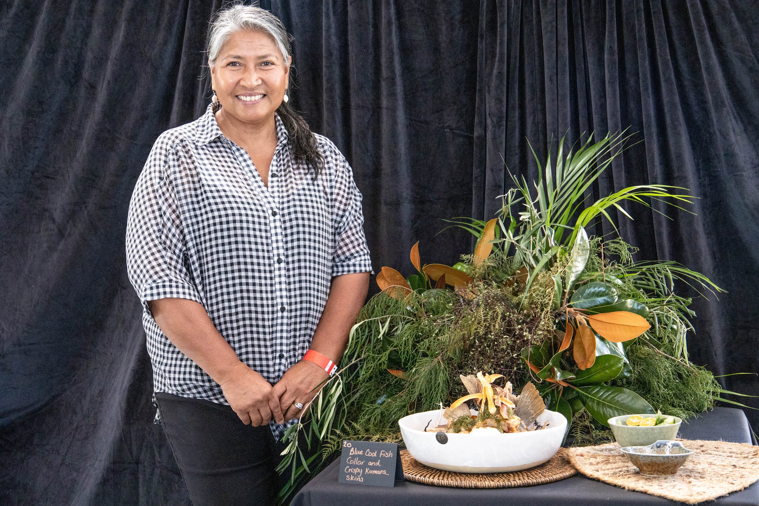 Woman with gray hair wearing a black-and-white checkered shirt standing beside a table with a large green plant arrangement and a white seafood display bowl, with a black background.