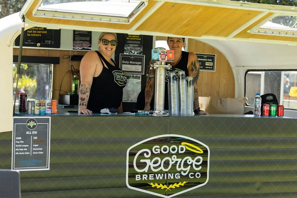 Two smiling women working at a mobile brewery stand labeled 'Good George Brewing Co', serving drinks and surrounded by cans, bottles, and cups, under a yellow canopy.