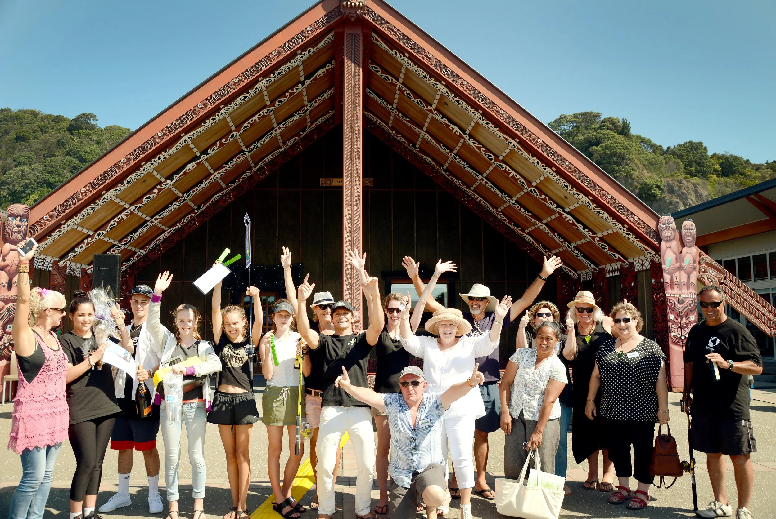 Group of smiling people, including children and adults, posing in front of a traditional Polynesian building with wooden carvings and a steeply pitched roof, outdoors on a sunny day.