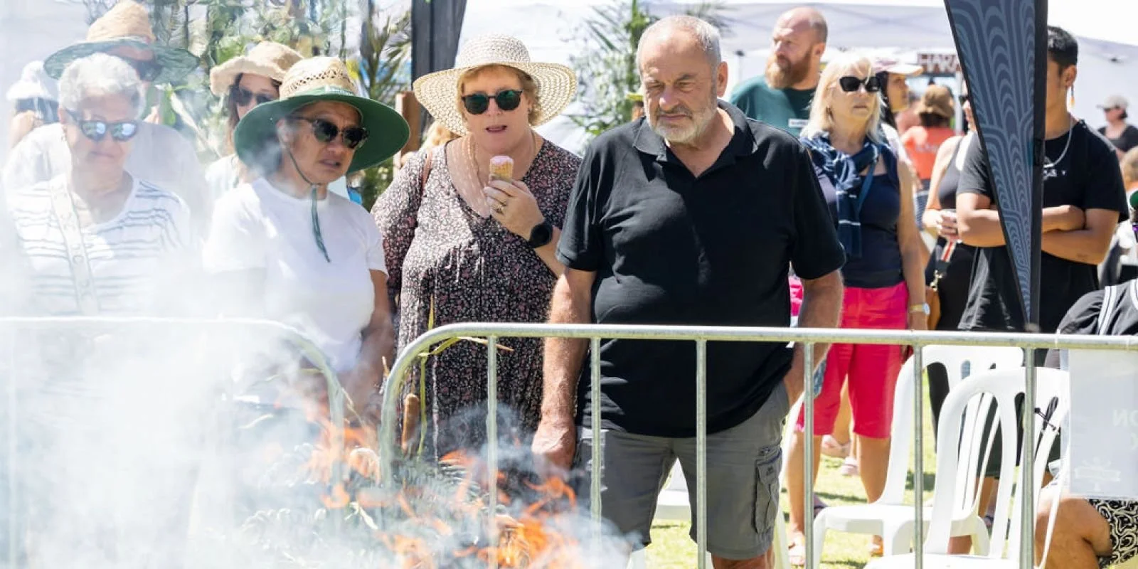 People at an outdoor event watching a barbecue grill with fire, some wearing hats and sunglasses, some holding snacks.