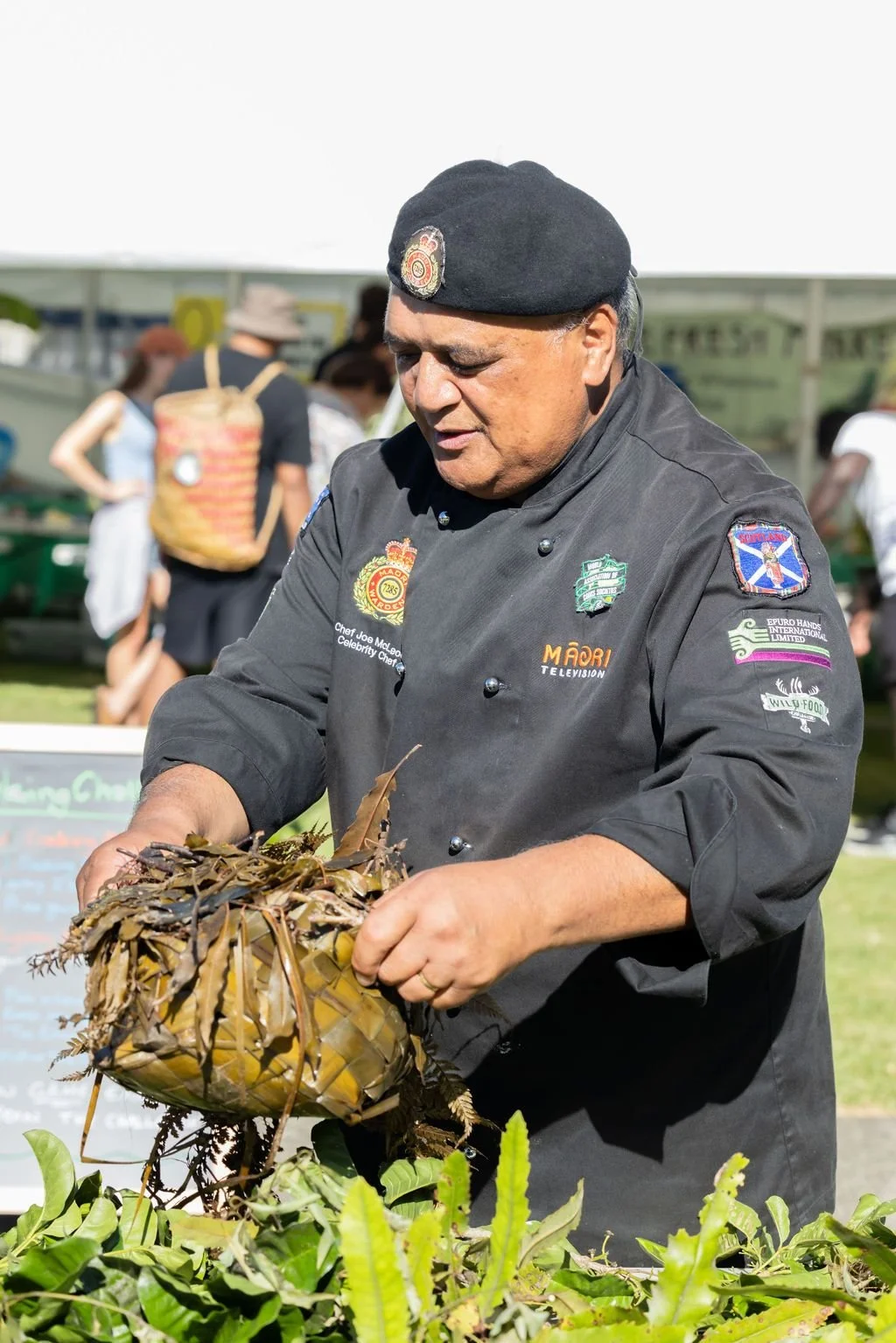 A chef in a black uniform and black beret displays a woven basket filled with leaves and natural materials at an outdoor event, with other people in the background.